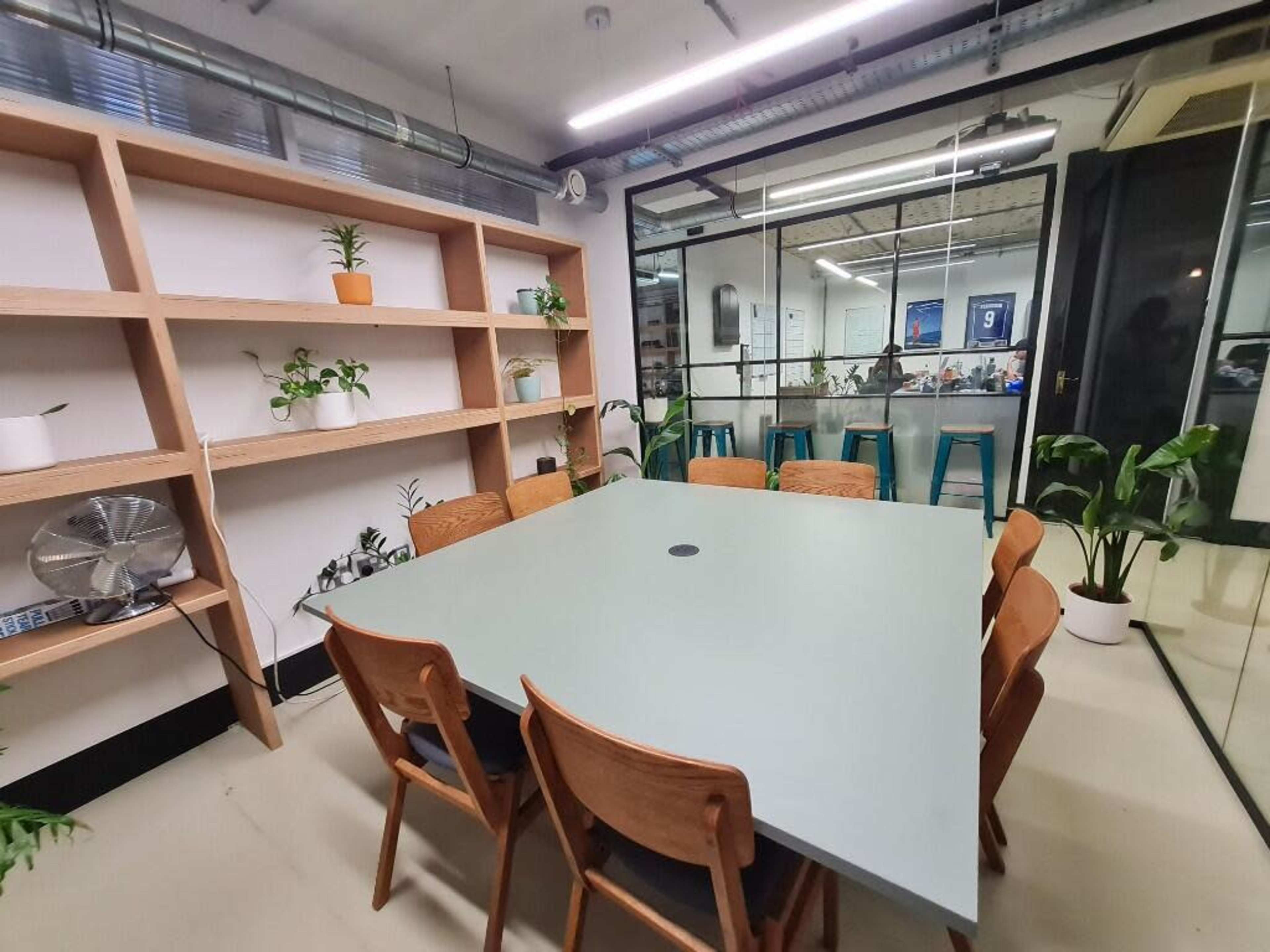 The image shows a modern meeting room with a large light green table surrounded by wooden chairs, and a plant shelf in the background.