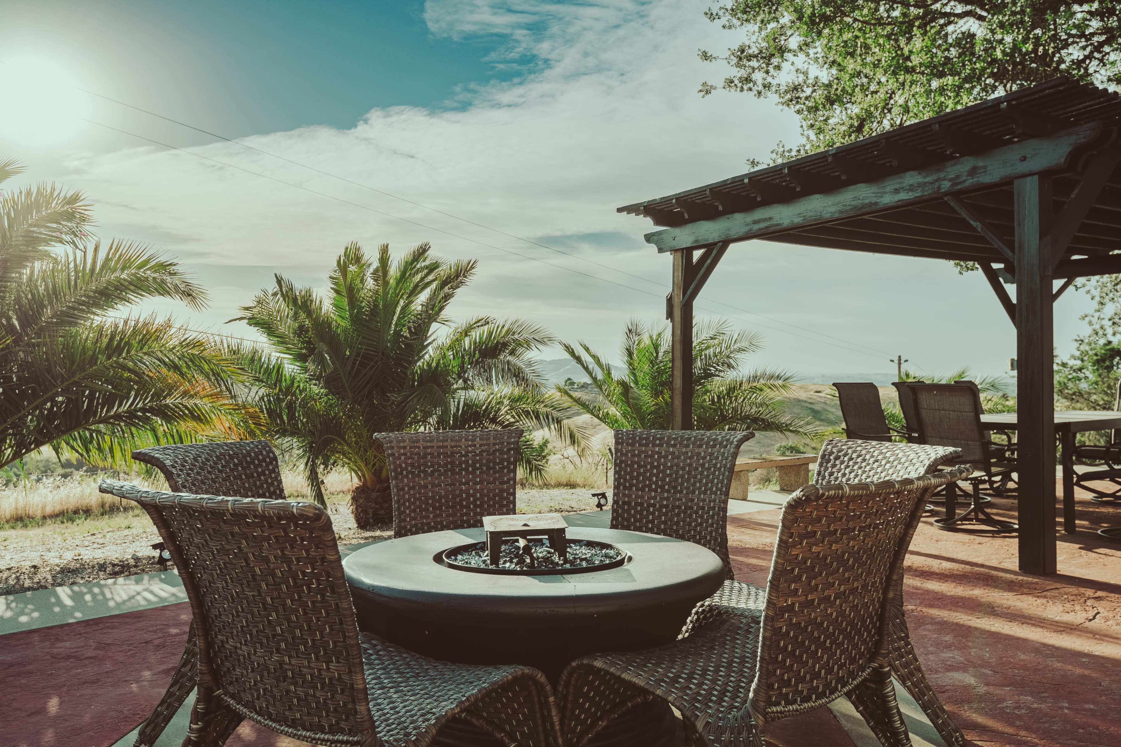 A circular table surrounded by woven chairs sits on a patio, with palm trees and a gazebo in the background under a clear sky.