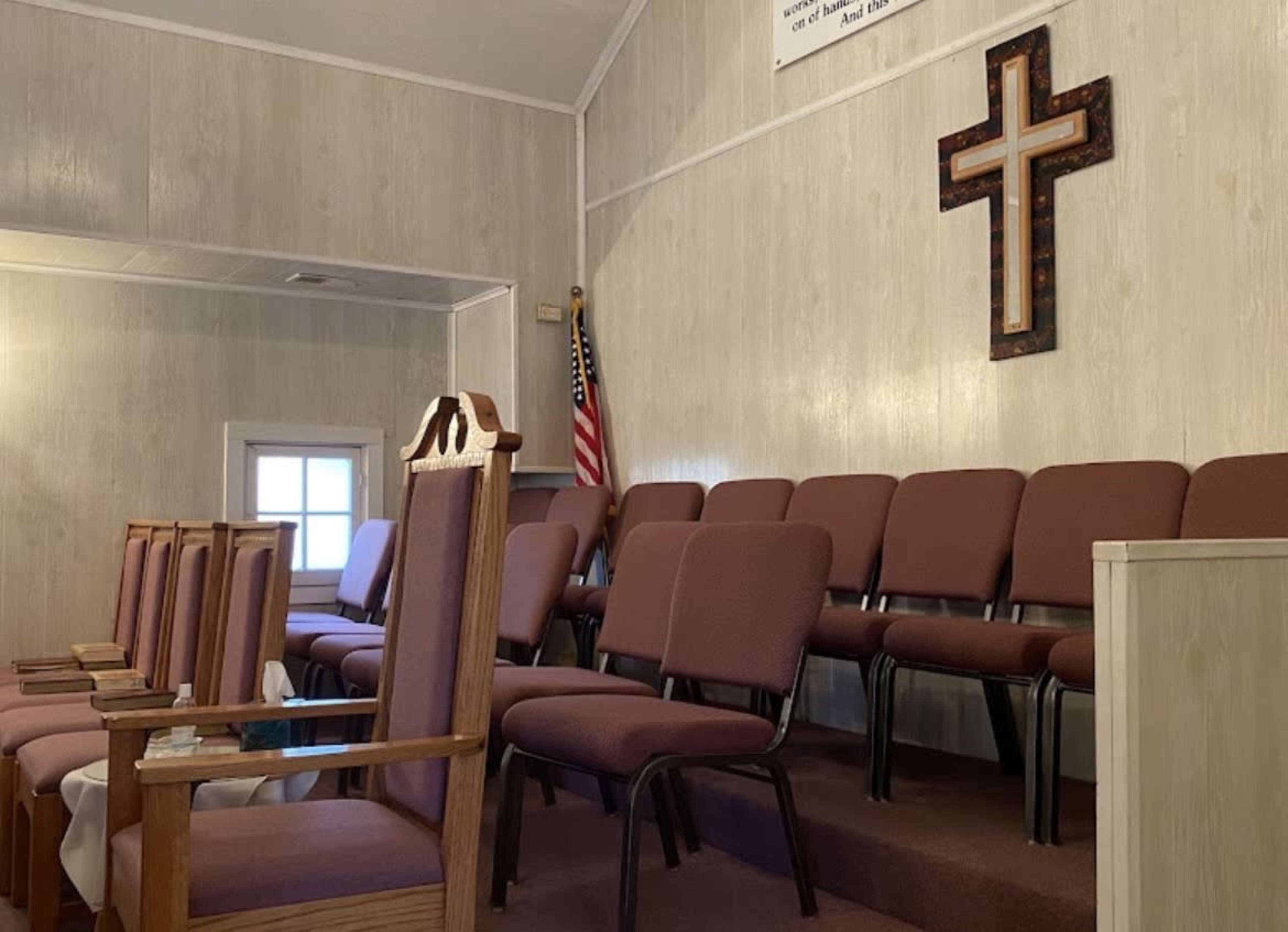 The interior of a church features empty maroon chairs arranged in rows, with a wooden cross and an American flag on the wall.