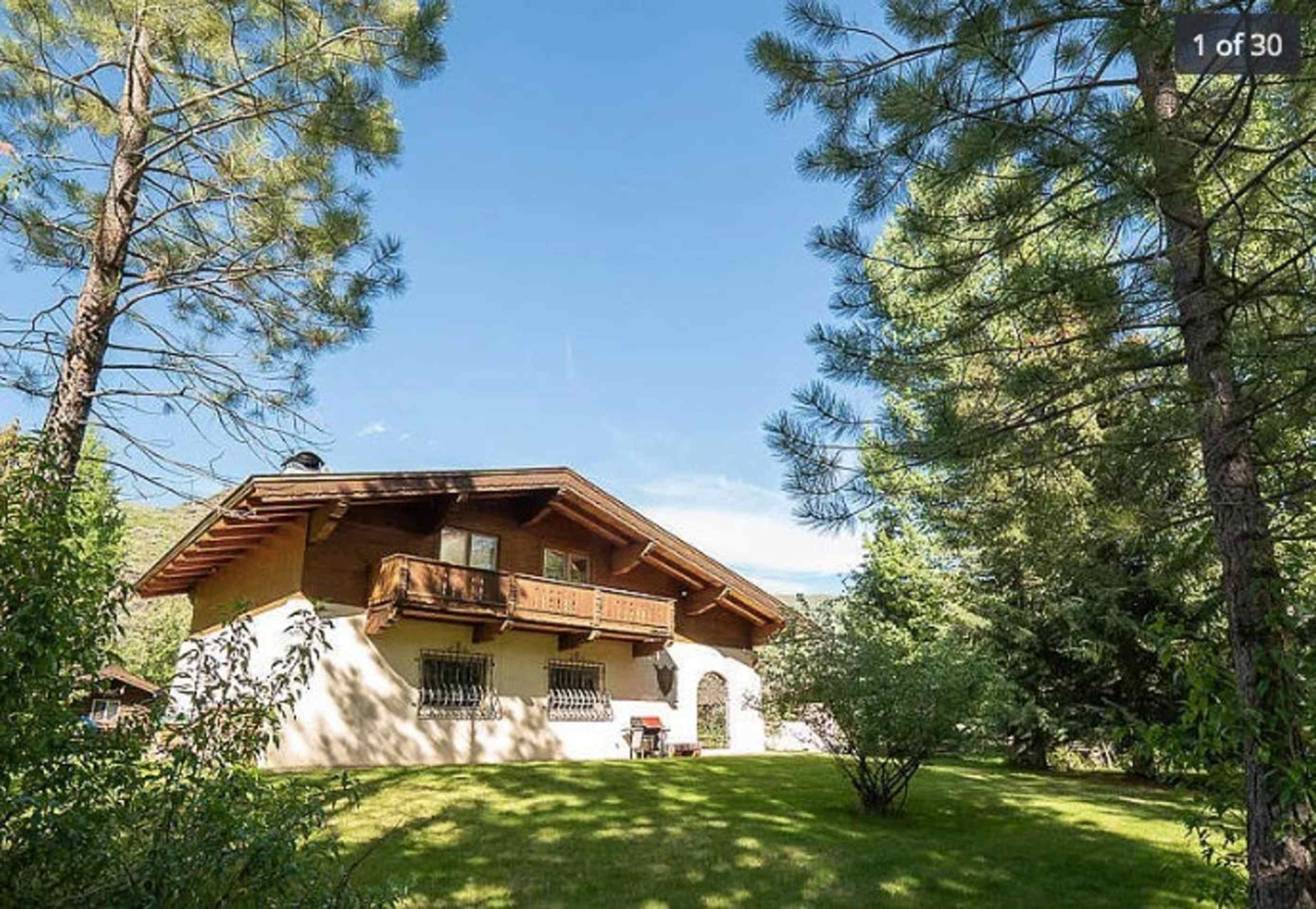 A large house with a wooden balcony sits amid green trees and grass under a clear blue sky.