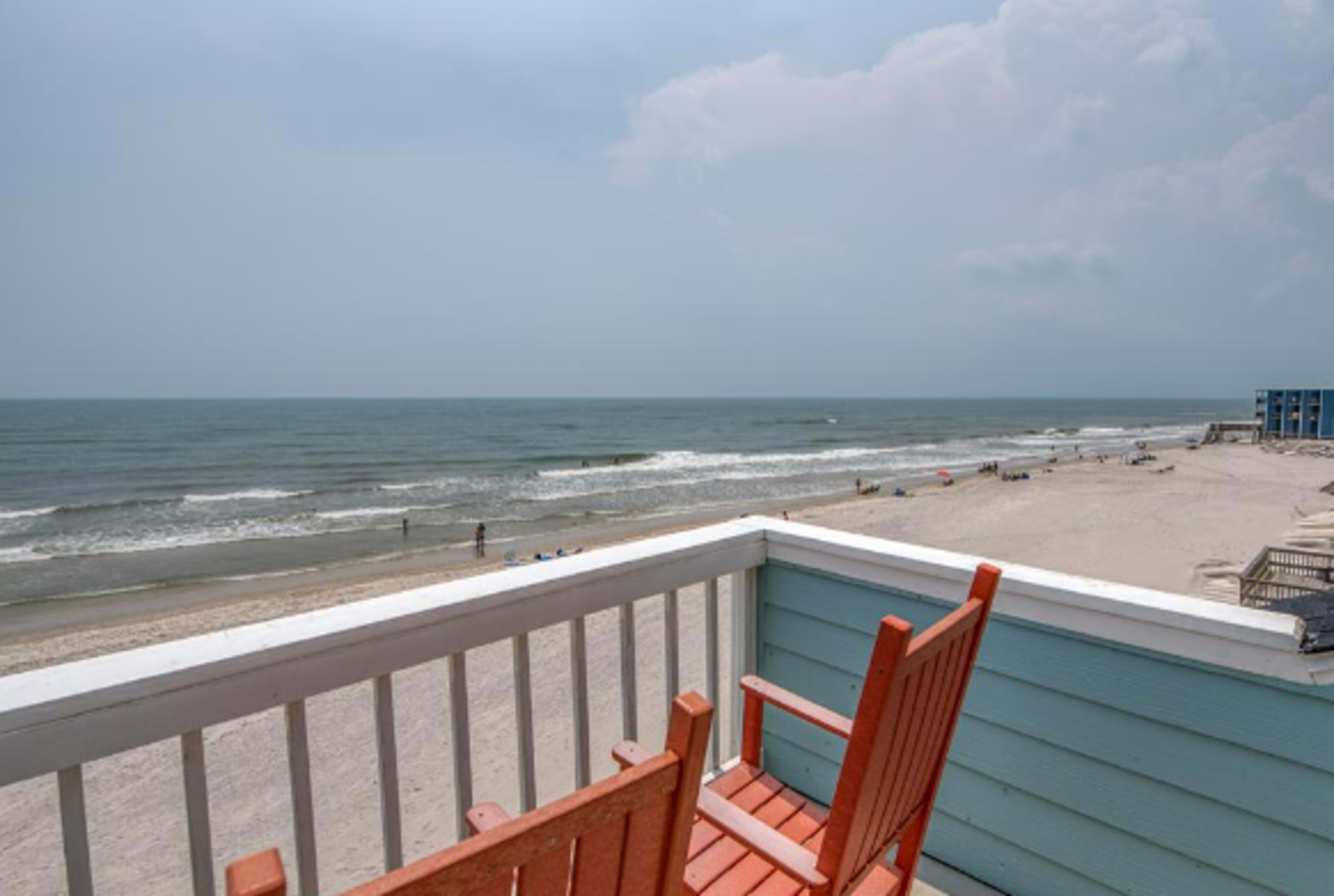 A balcony view overlooking a sandy beach and the ocean, with two orange chairs positioned at the edge.
