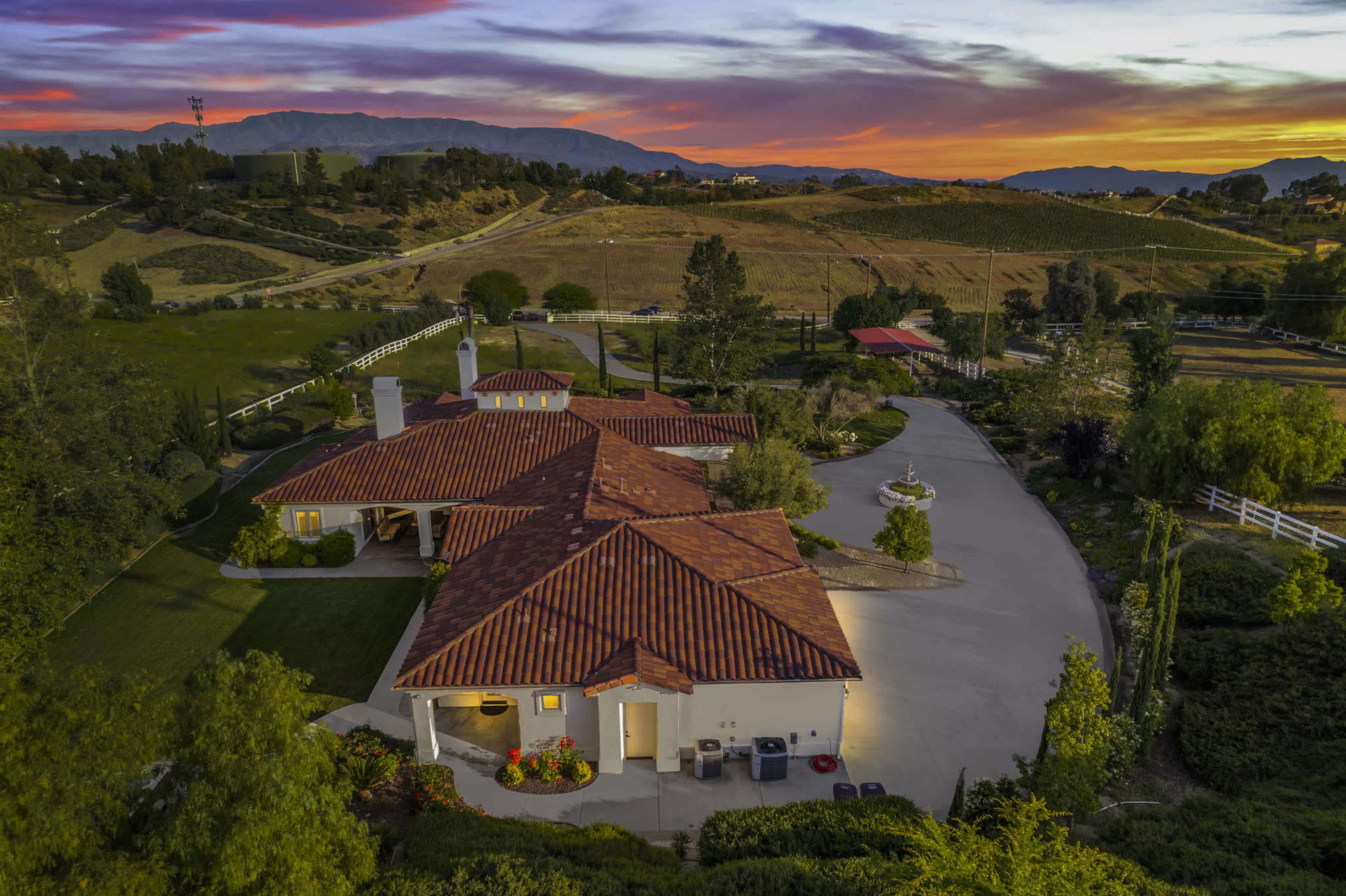 A large residential property with a tile-roofed house surrounded by green lawns and trees, set against a sunset backdrop over distant mountains.