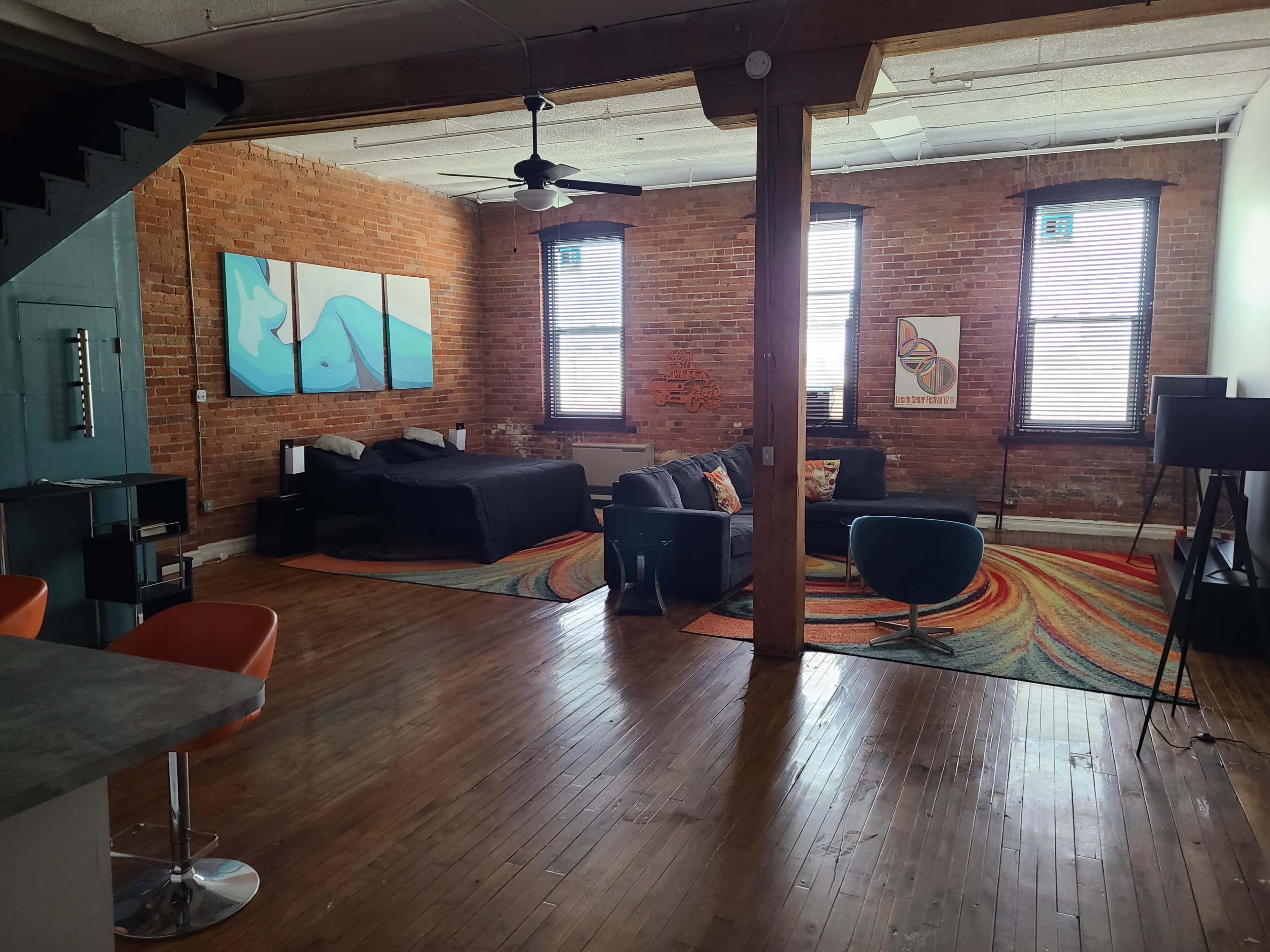 A spacious loft-style living area with exposed brick walls, large windows, a black bed, and modern furniture arranged on a colorful patterned rug.