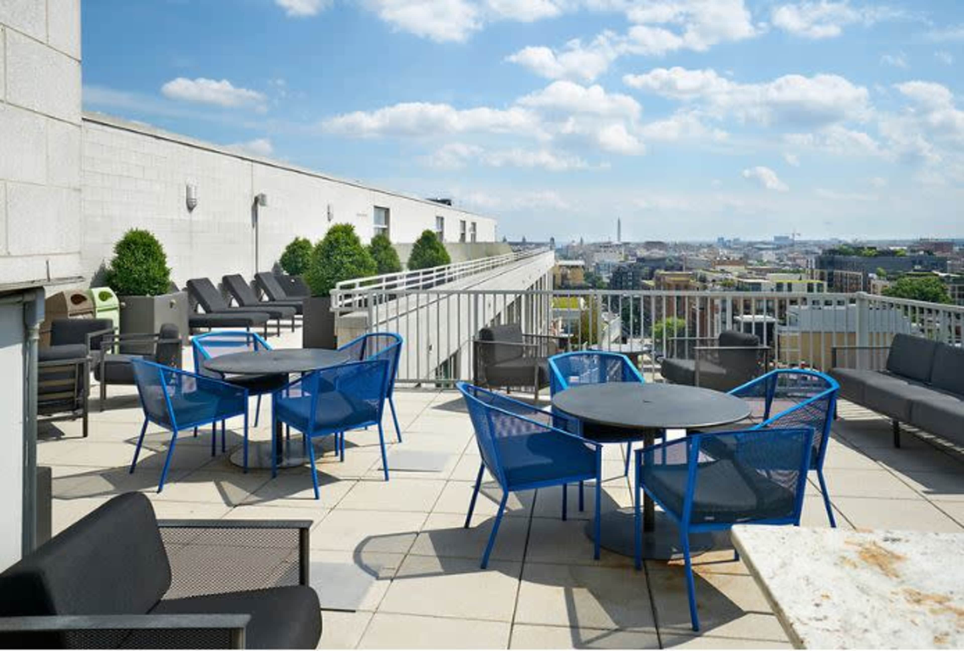 A rooftop terrace with blue chairs and tables, surrounded by a city skyline under a partly cloudy sky.