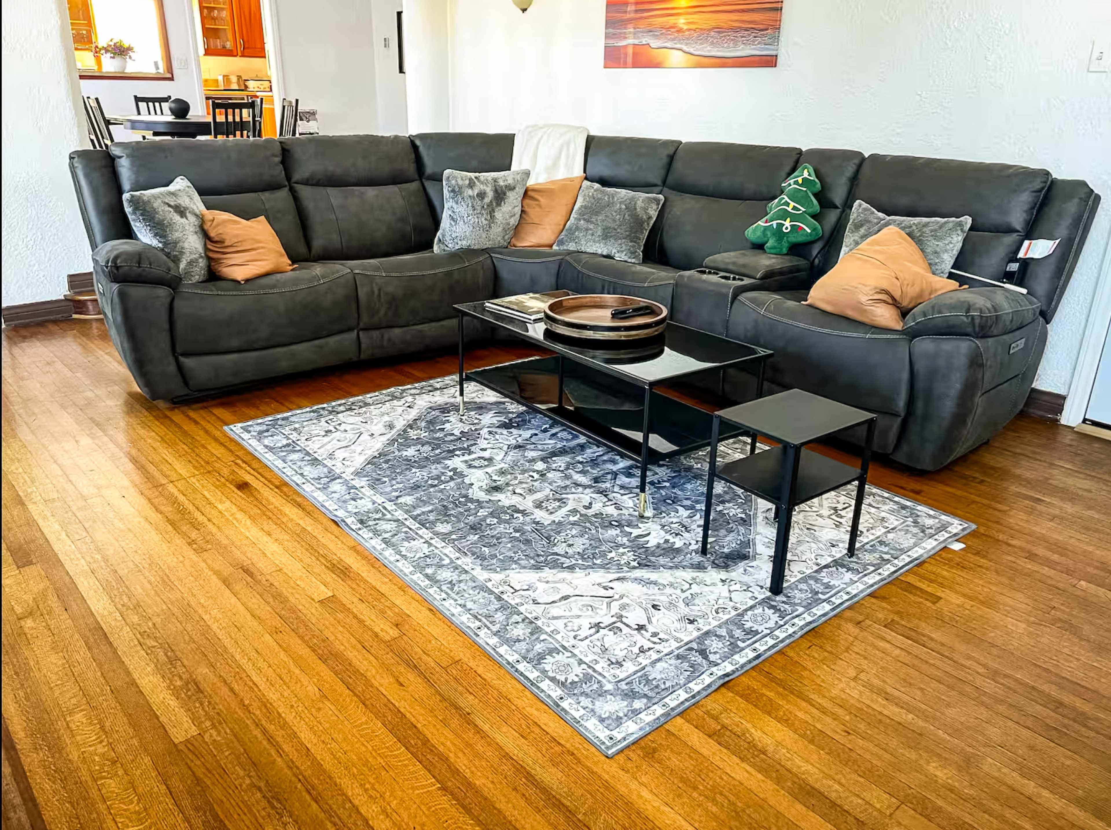 The image shows a dark gray sectional sofa adorned with decorative pillows, a glass coffee table, and a patterned rug on a hardwood floor in a well-lit living space.