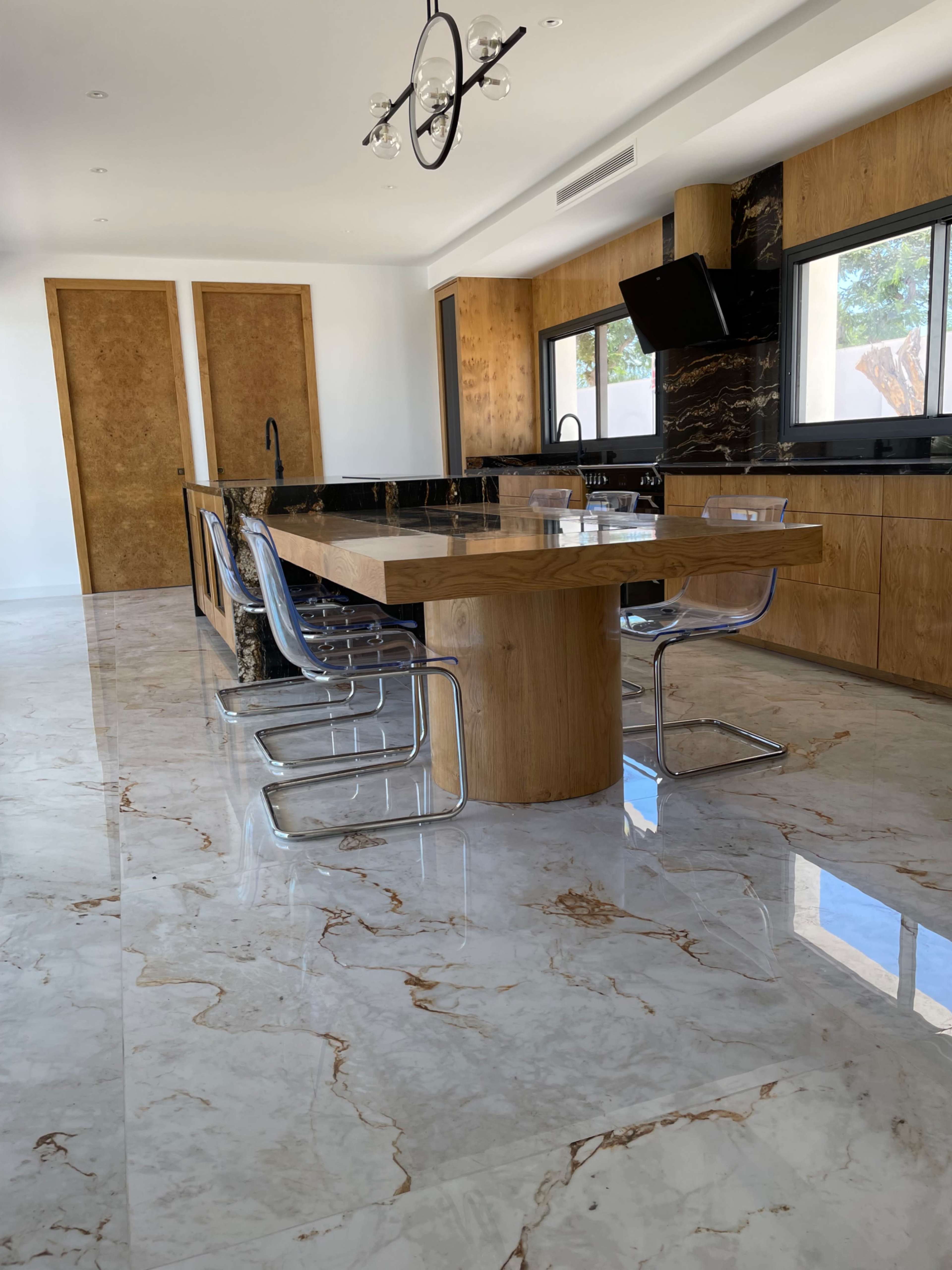 A modern kitchen features a marble floor, a wooden dining table surrounded by chrome chairs, and large windows.