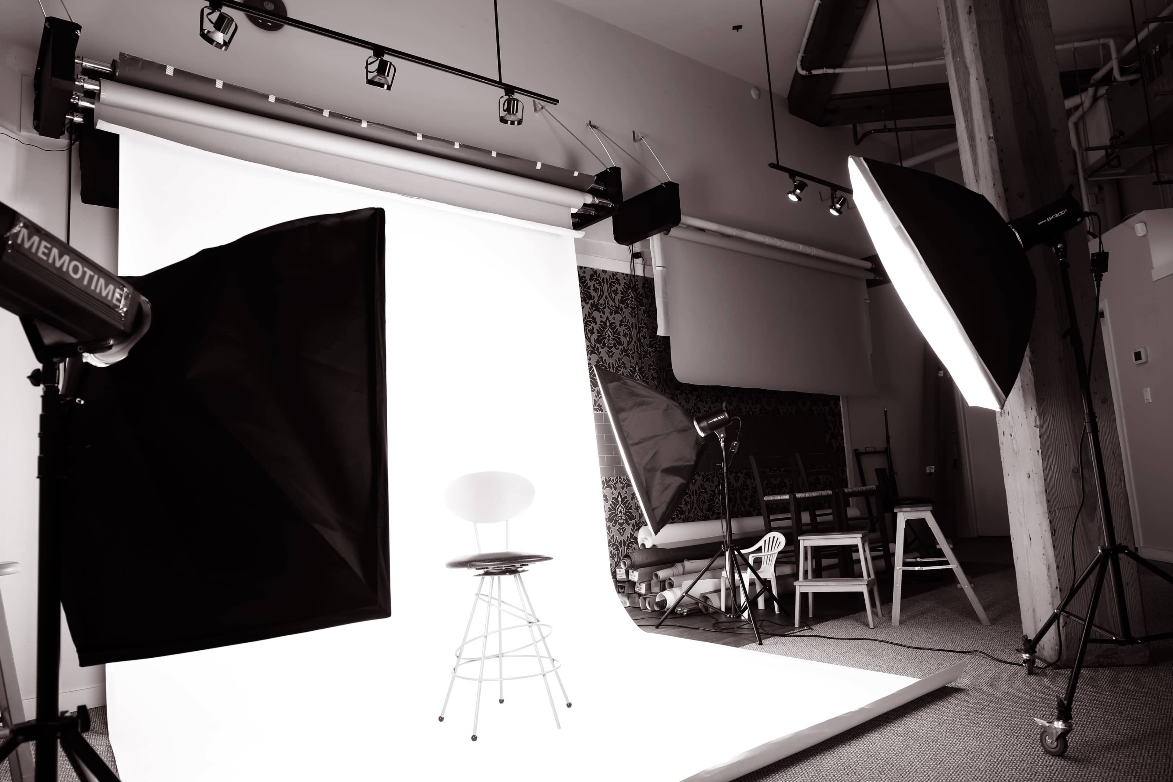 A photography studio features a simple backdrop with lighting equipment and an empty stool positioned for a photoshoot.