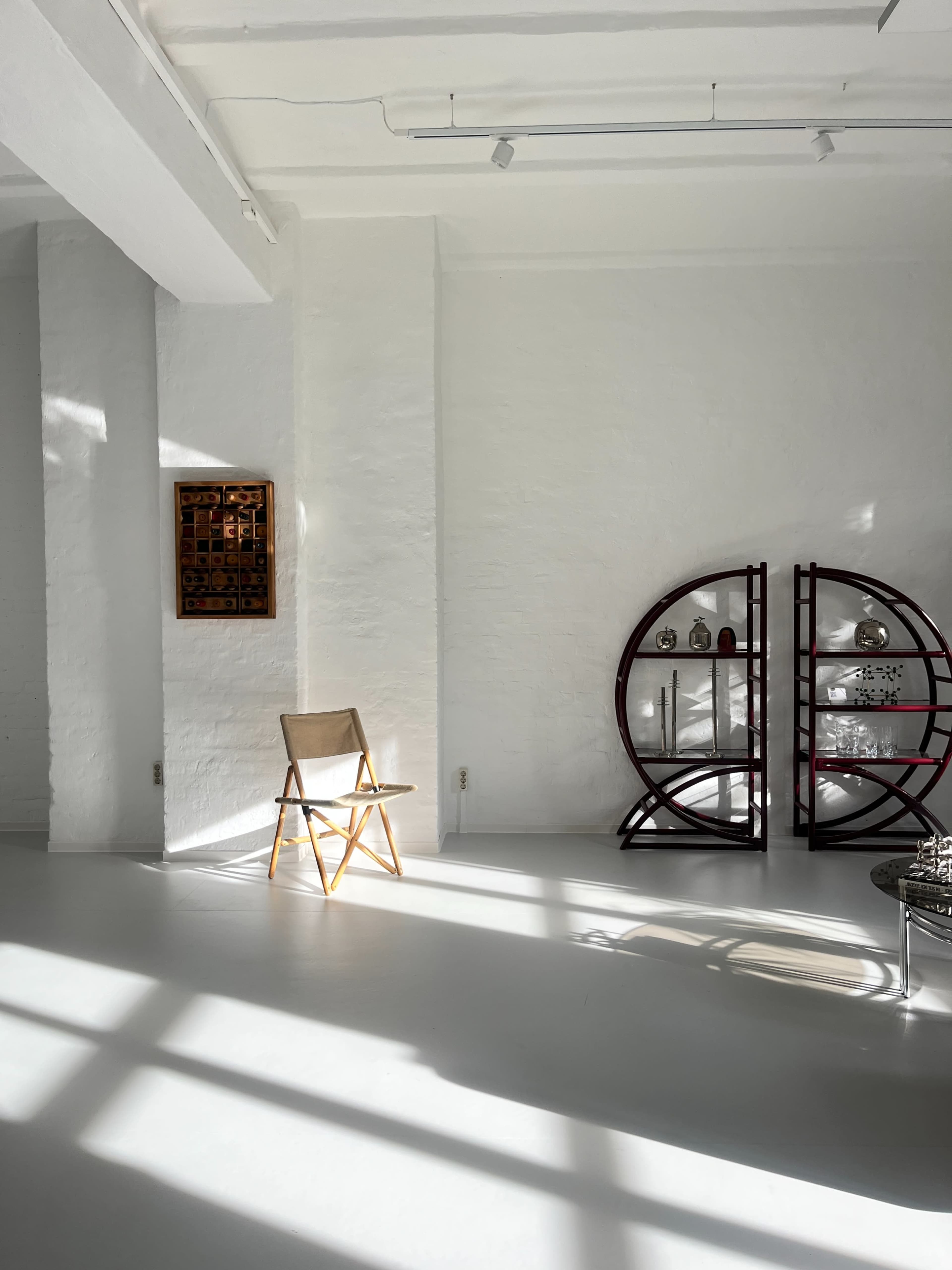 The image shows a bright, minimalist room with a wooden chair and a curved shelf displaying decorative items against white walls.