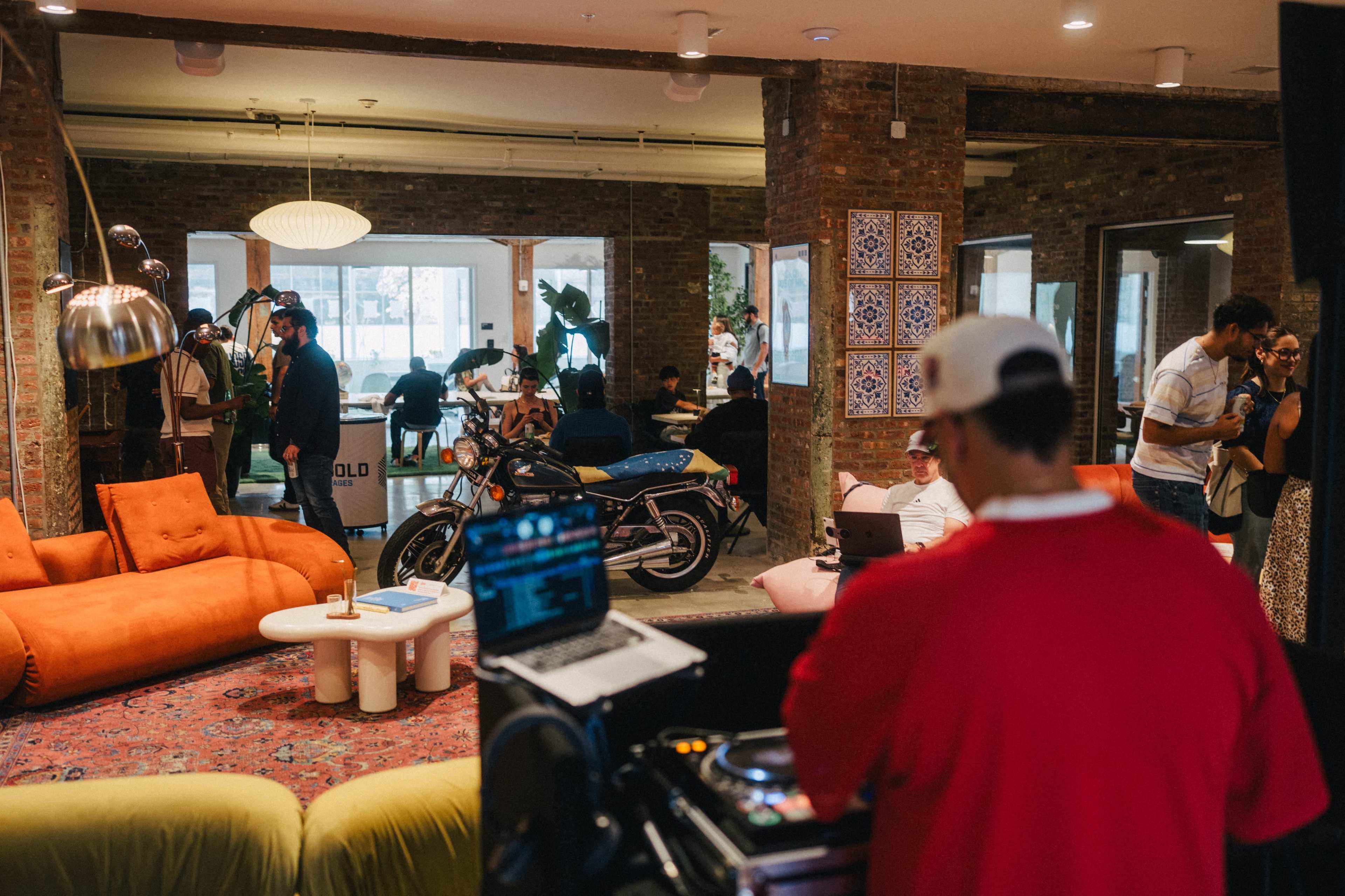 A lively coworking space features a DJ setup in the foreground, colorful couches, and people engaging in conversation and work throughout the room.