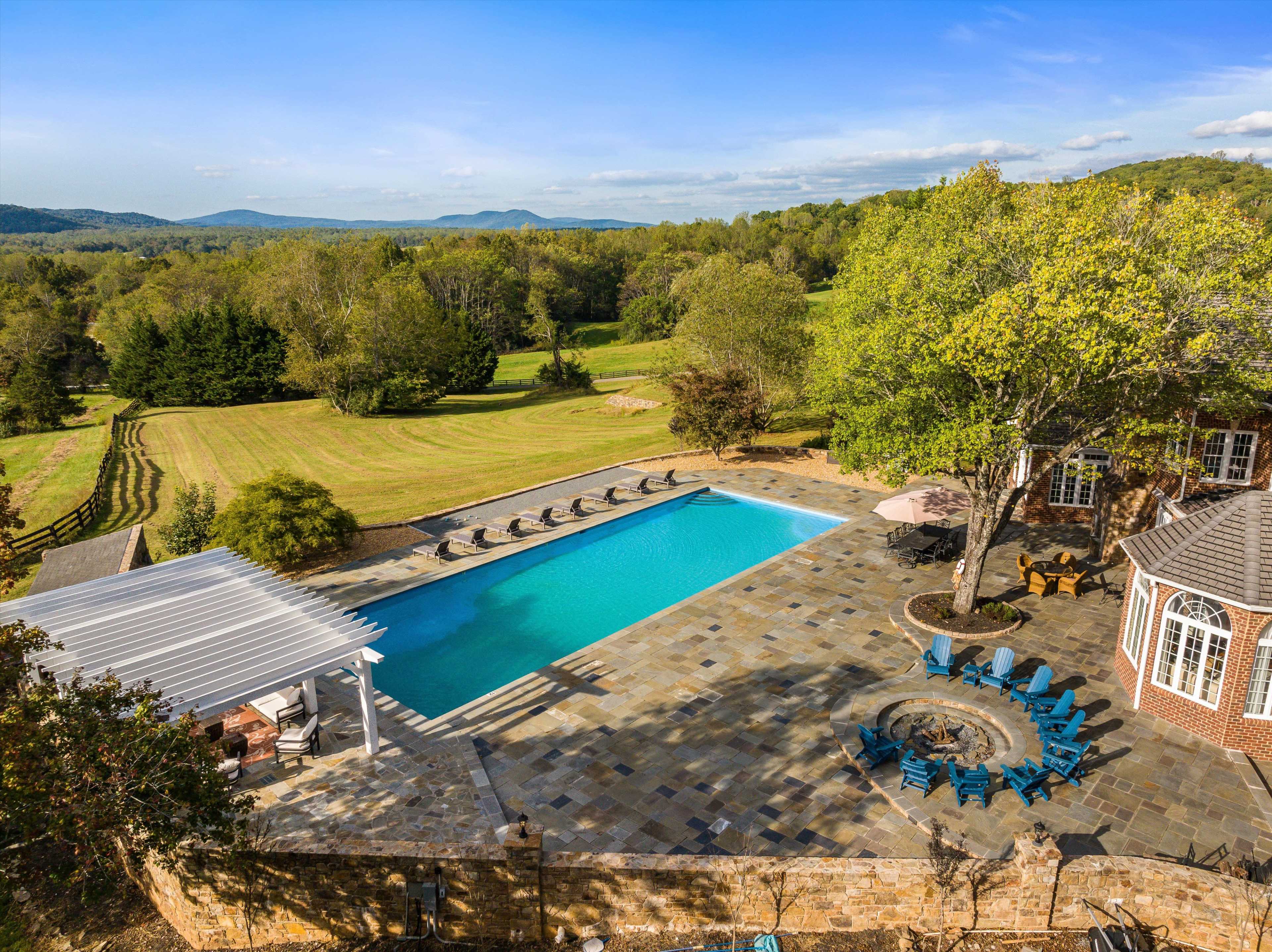 The image shows a large outdoor pool surrounded by stone paving, lounge chairs, and a fire pit, set against a scenic landscape of trees and rolling hills.
