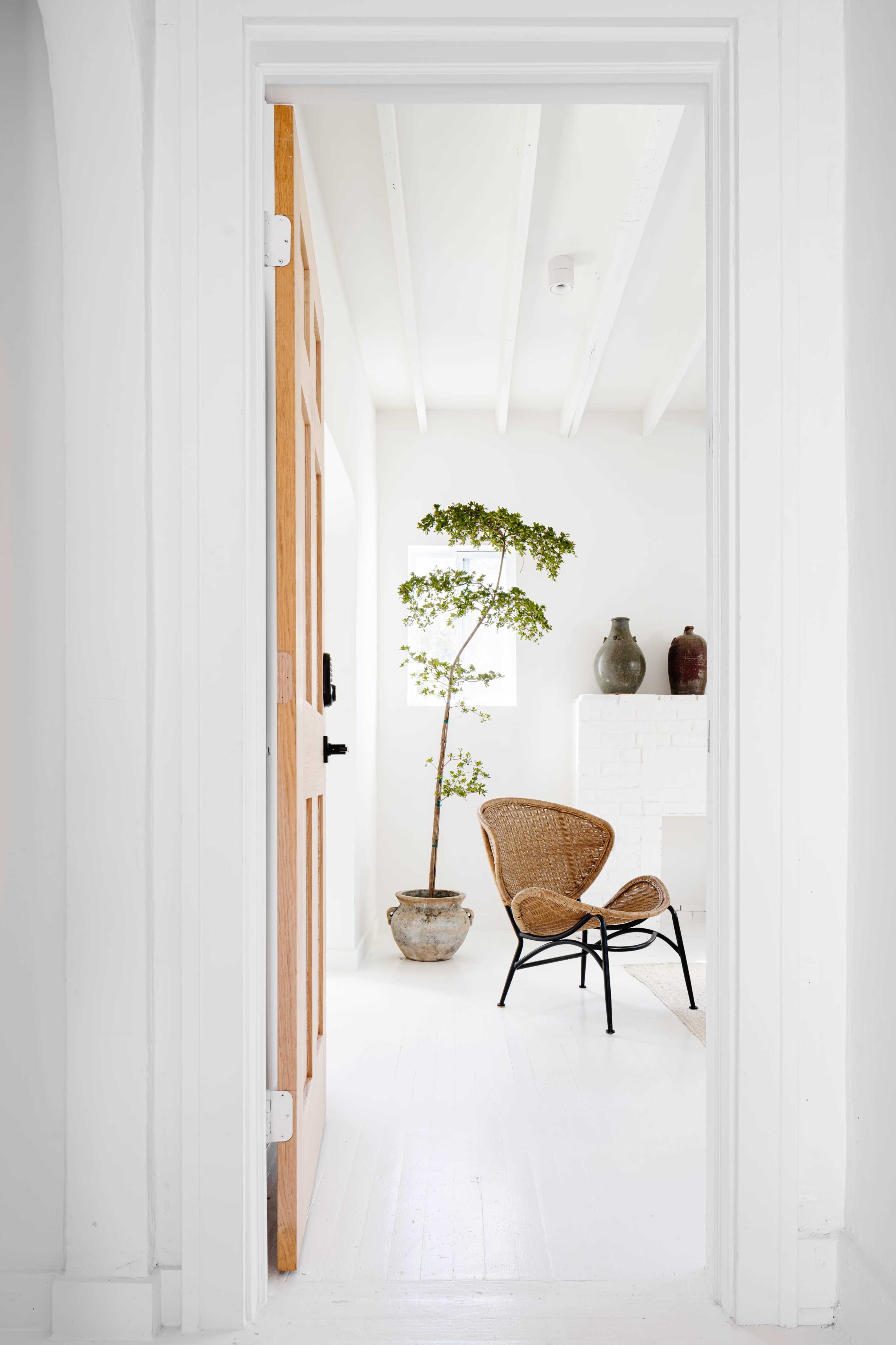 A wooden door opens to a bright room featuring a chair, a potted plant, and two decorative vases.