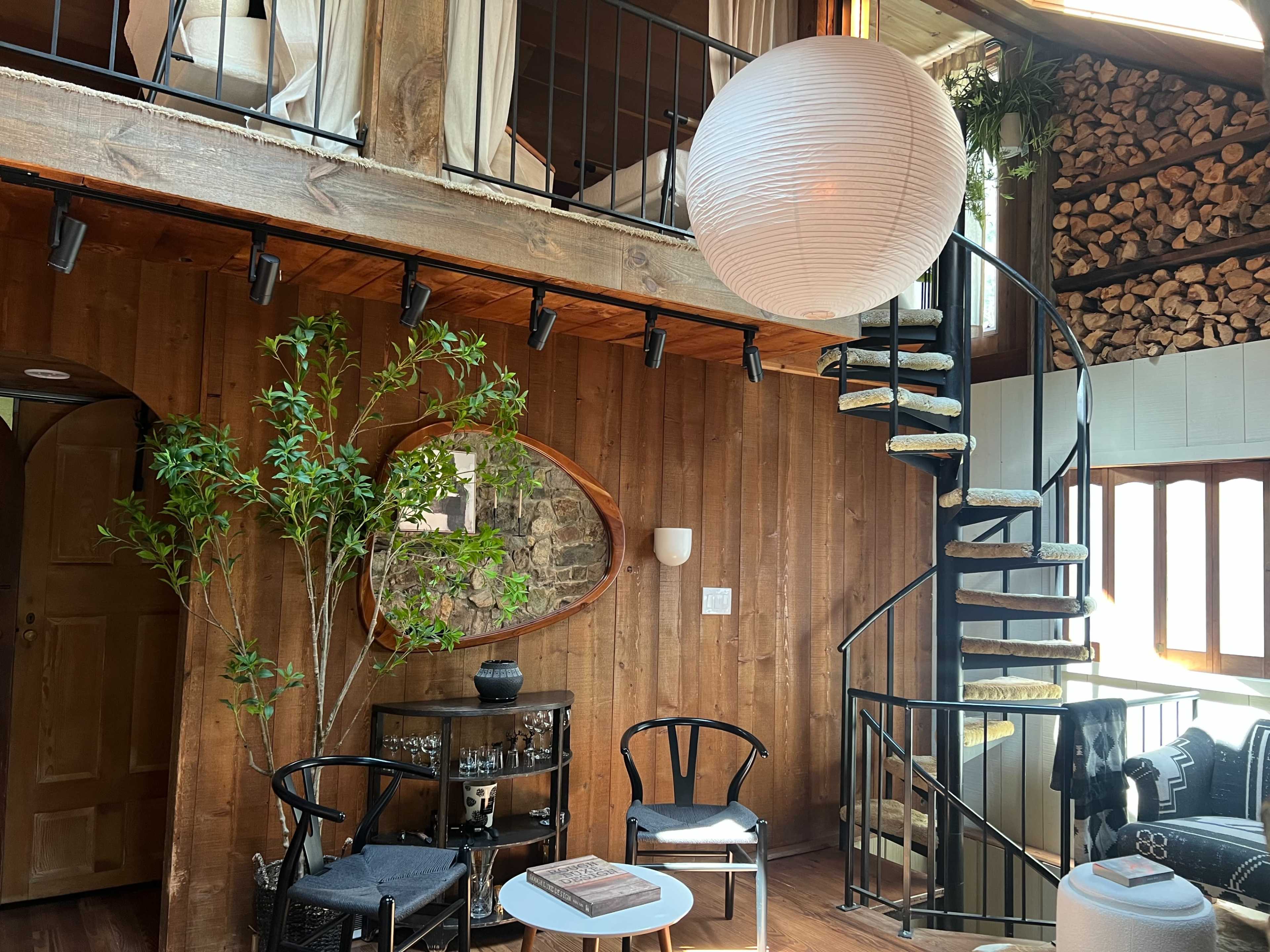 The image depicts a cozy room with wooden walls, a spiraling staircase, a large round pendant light, and a small seating area featuring a table and chairs.