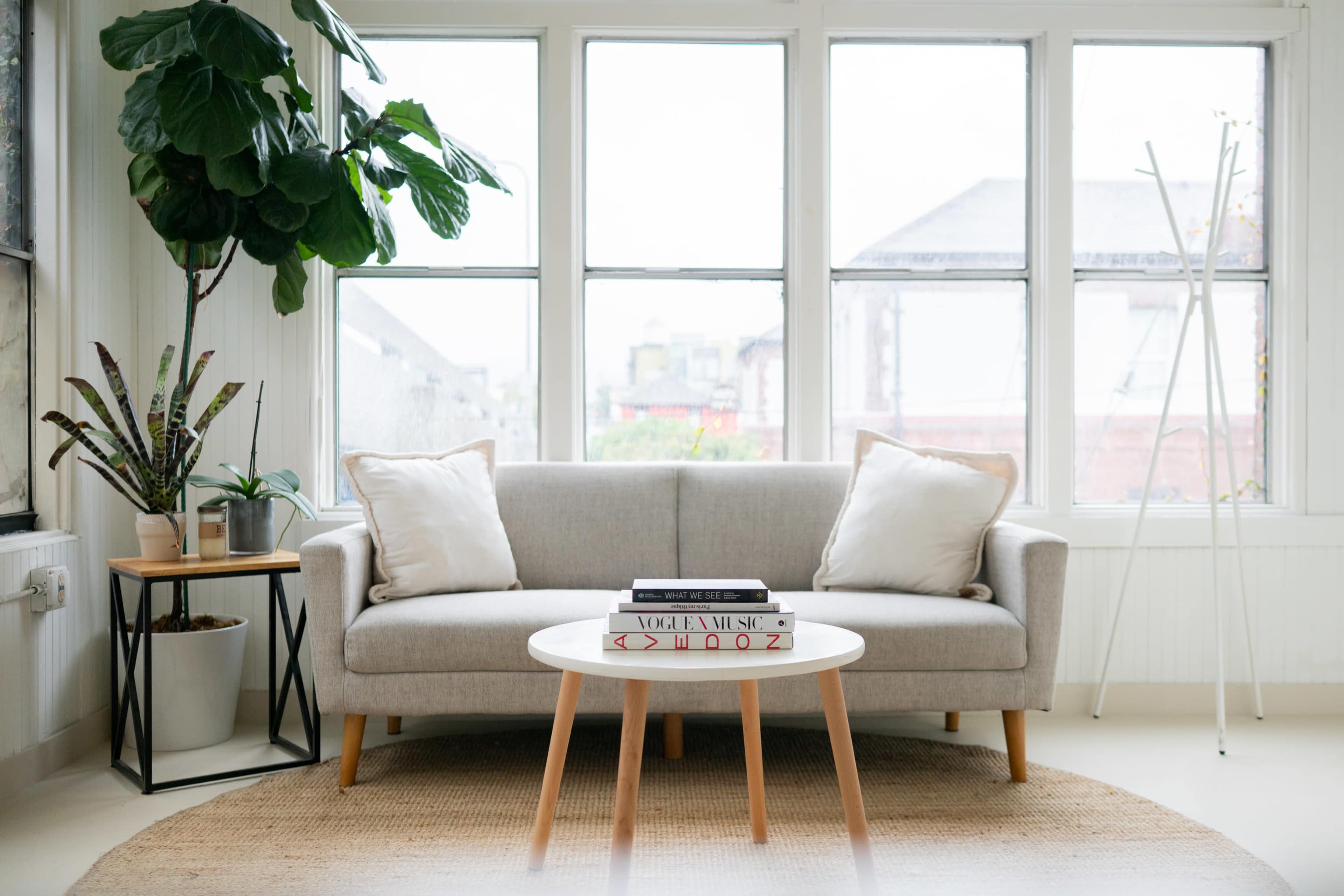 A light-filled living room features a gray sofa with pillows, a round coffee table, and potted plants, surrounded by large windows.