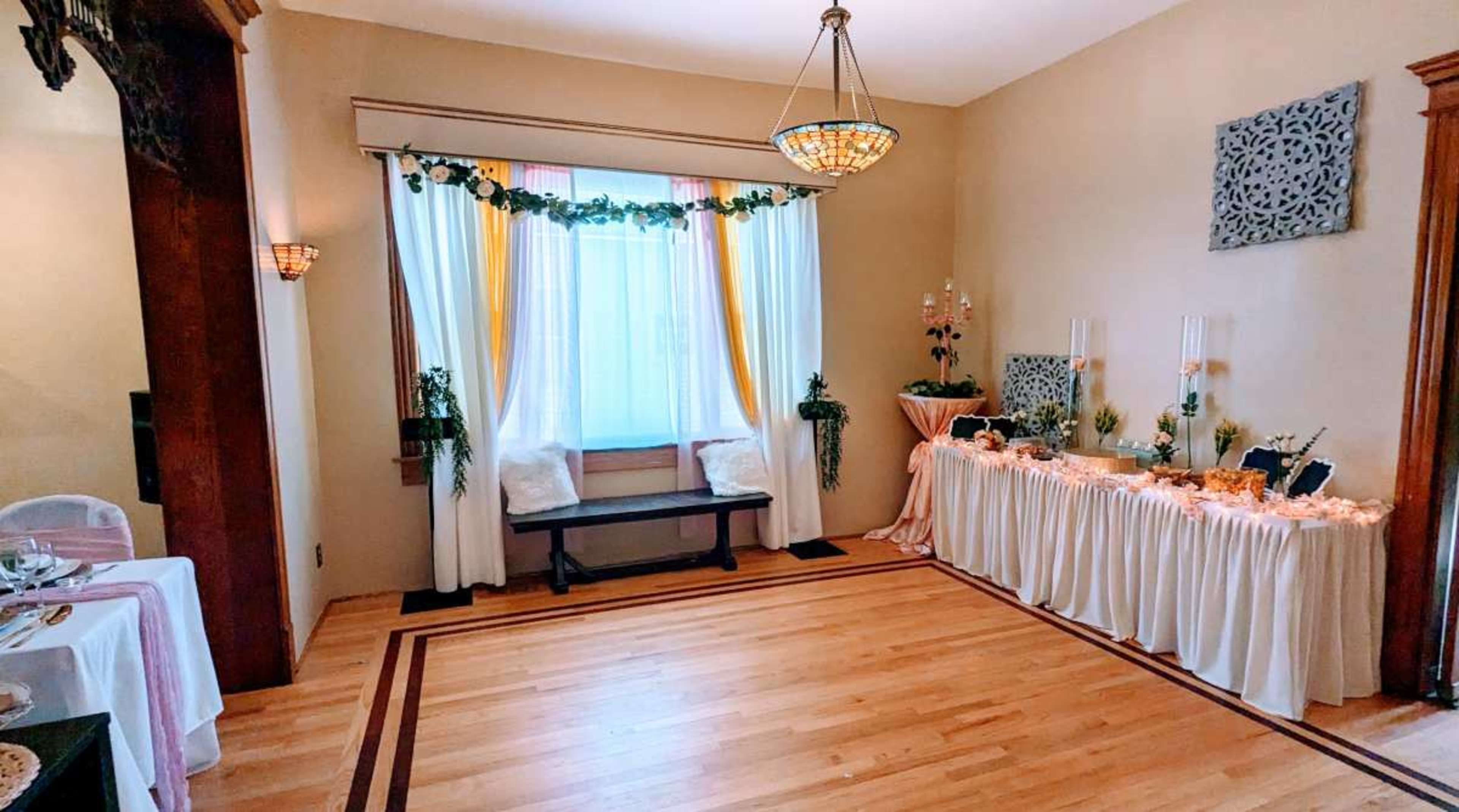 The room features a window adorned with curtains, a wooden bench beneath it, and a decorative table set with tablecloths and various floral arrangements.