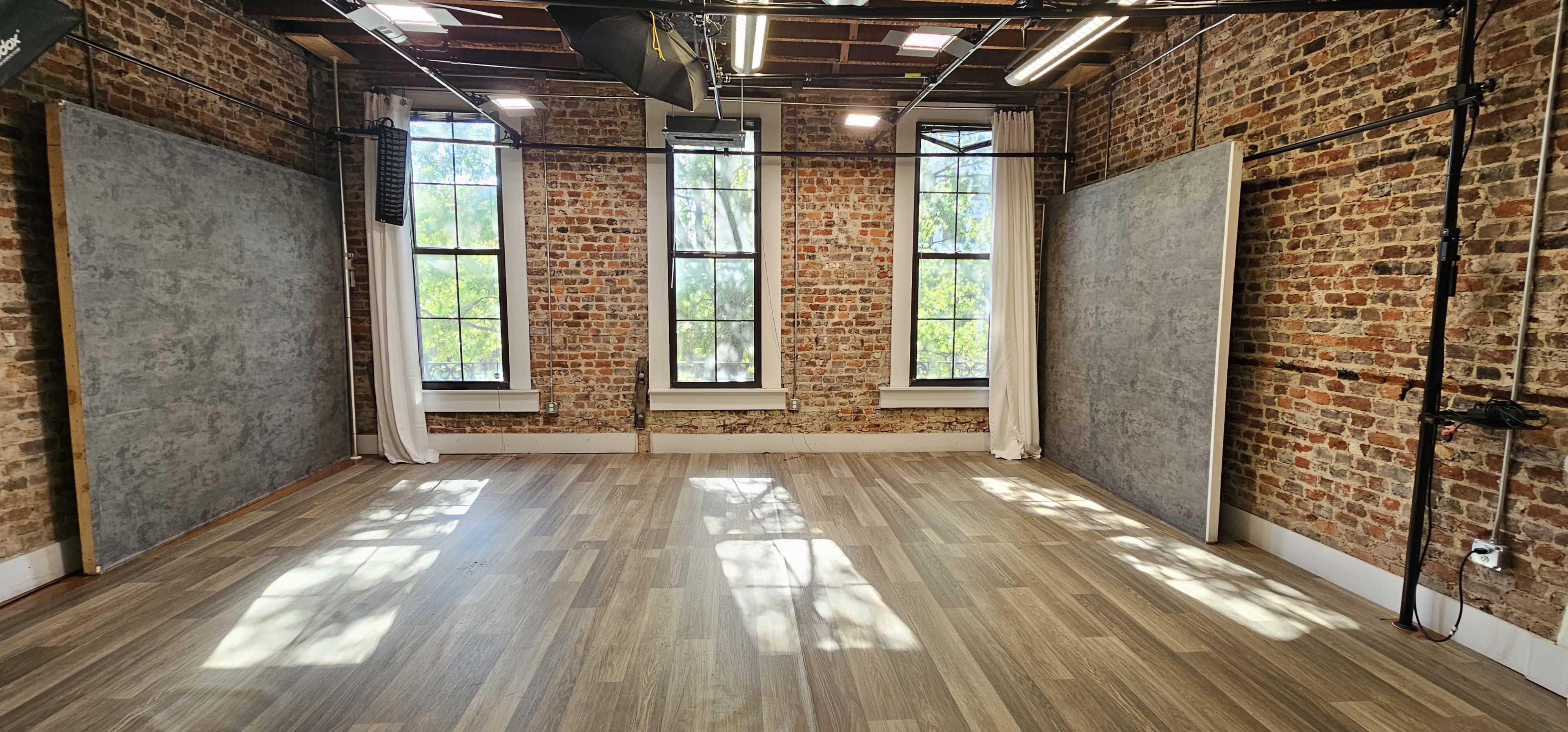 The image shows an empty studio space with exposed brick walls, large windows allowing natural light, and two gray backdrop panels on either side.
