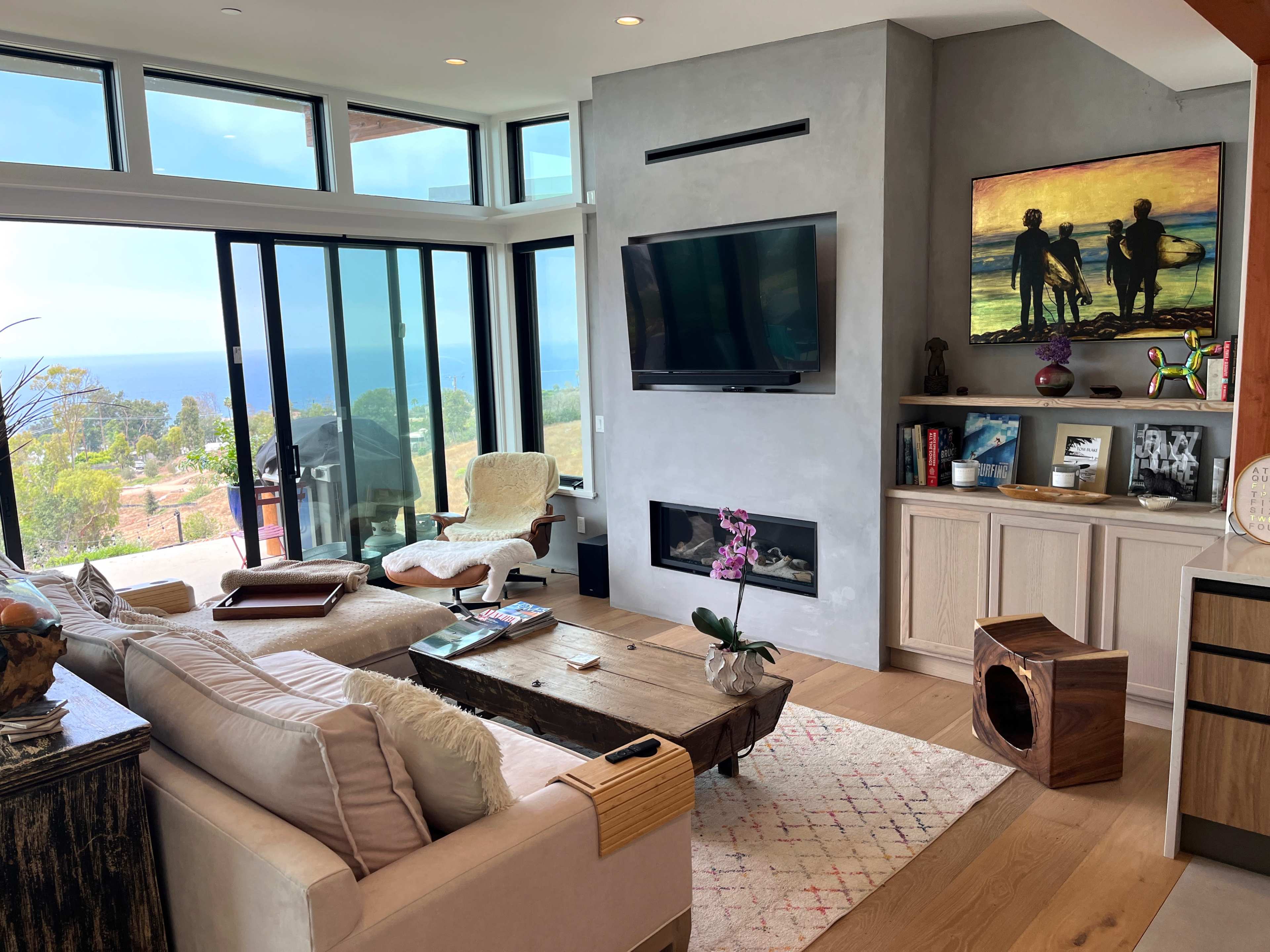 The image shows a modern living room with large windows revealing a view of the ocean, featuring a comfortable sofa, a coffee table, a TV, and decorative artwork on the walls.