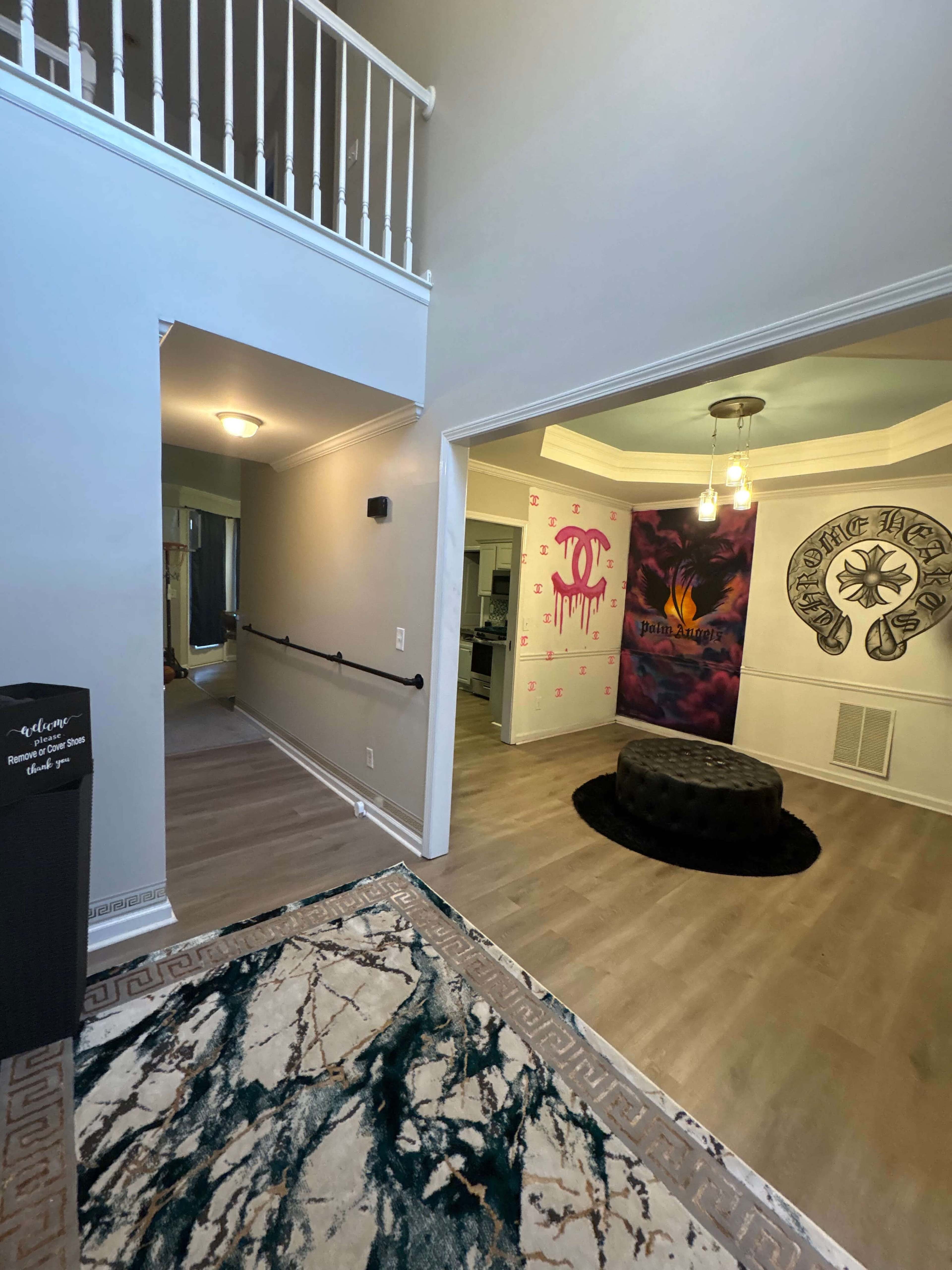 The image shows a spacious entryway leading into a living area, featuring a circular black ottoman, a patterned rug, and walls adorned with colorful artwork.
