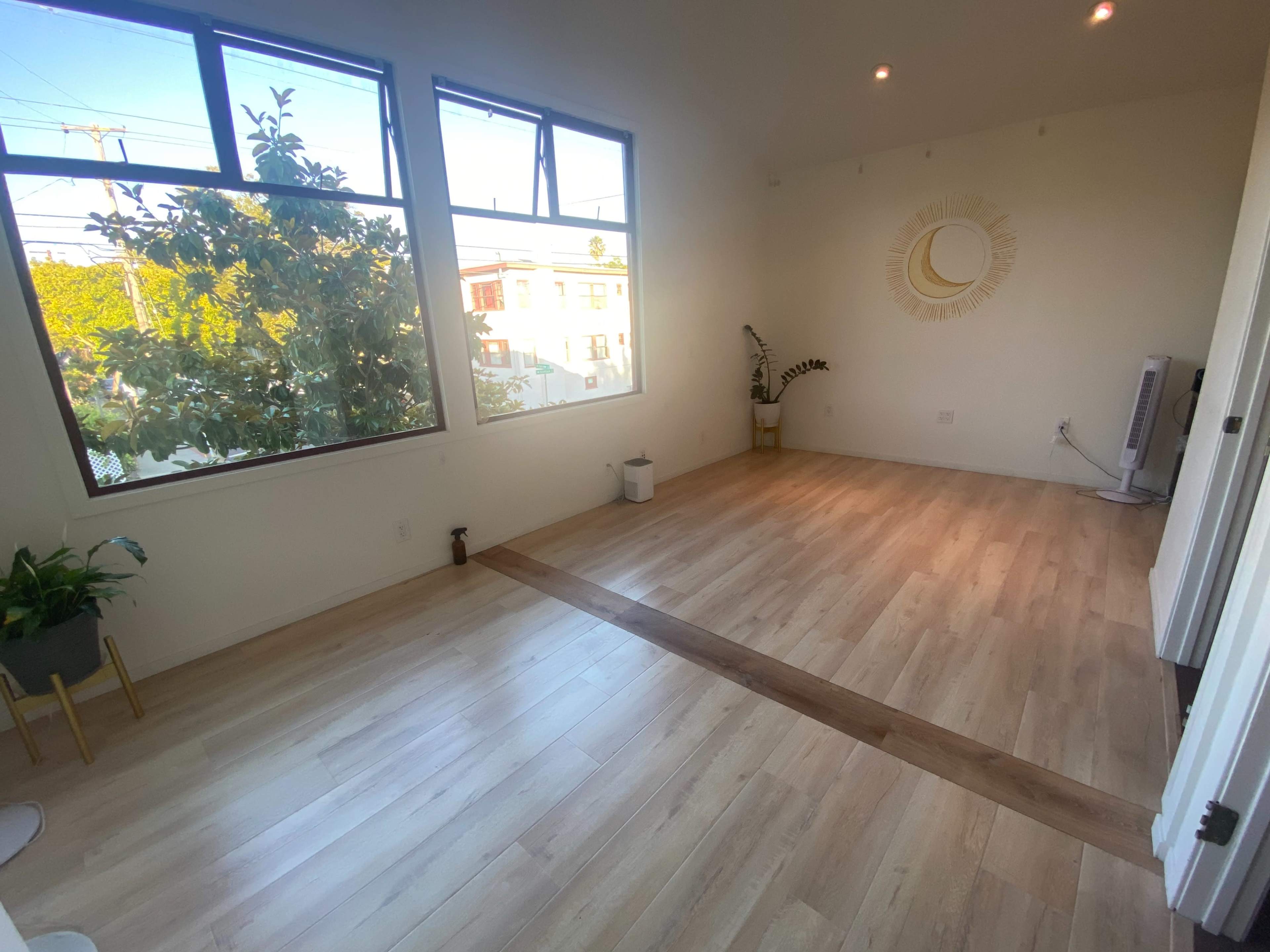 A bright, empty room with large windows, wooden flooring, and a circular decorative piece on the wall.