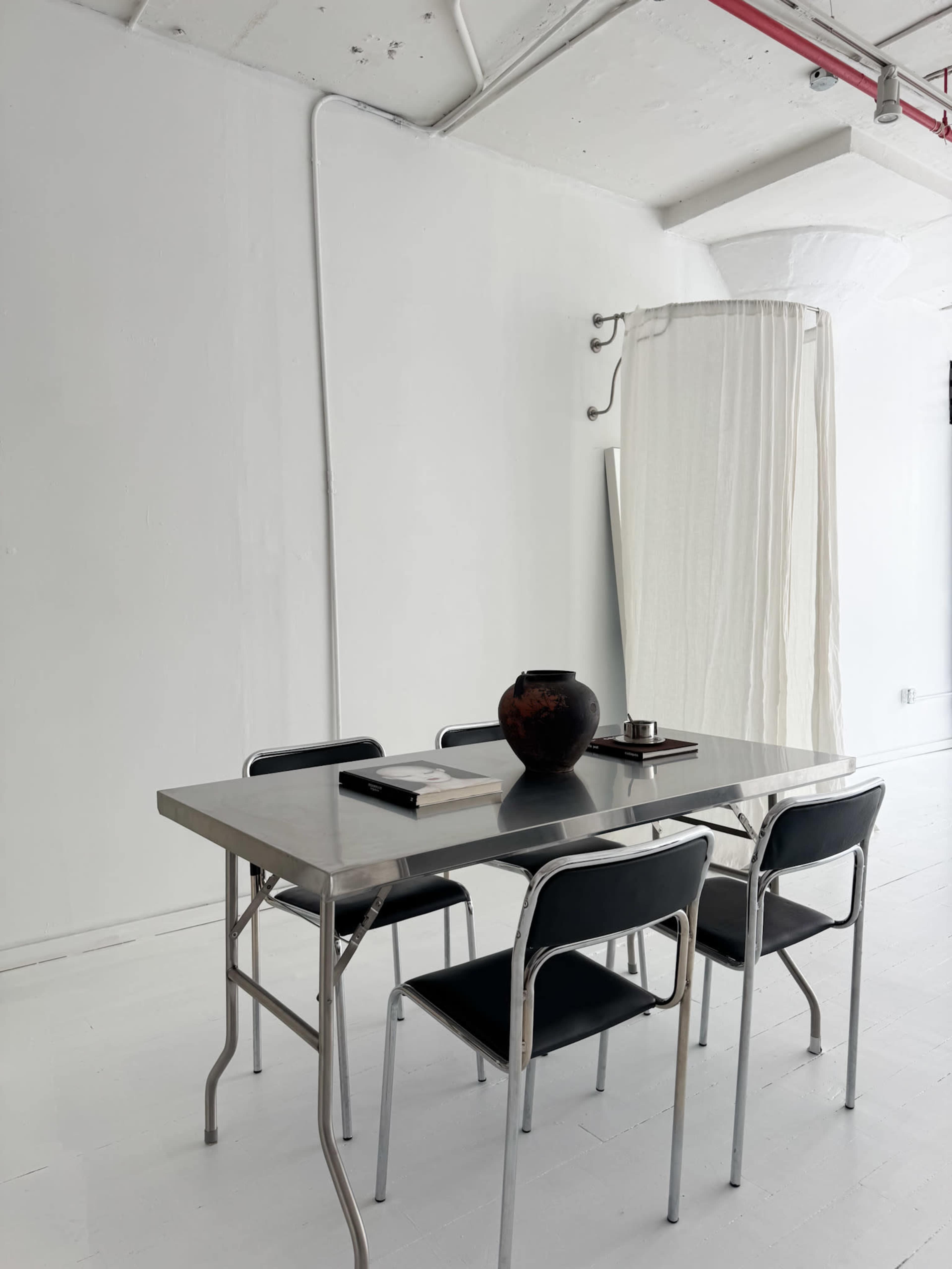 A minimalist dining table with four black chairs is positioned in a bright, empty room featuring white walls and light-colored flooring.