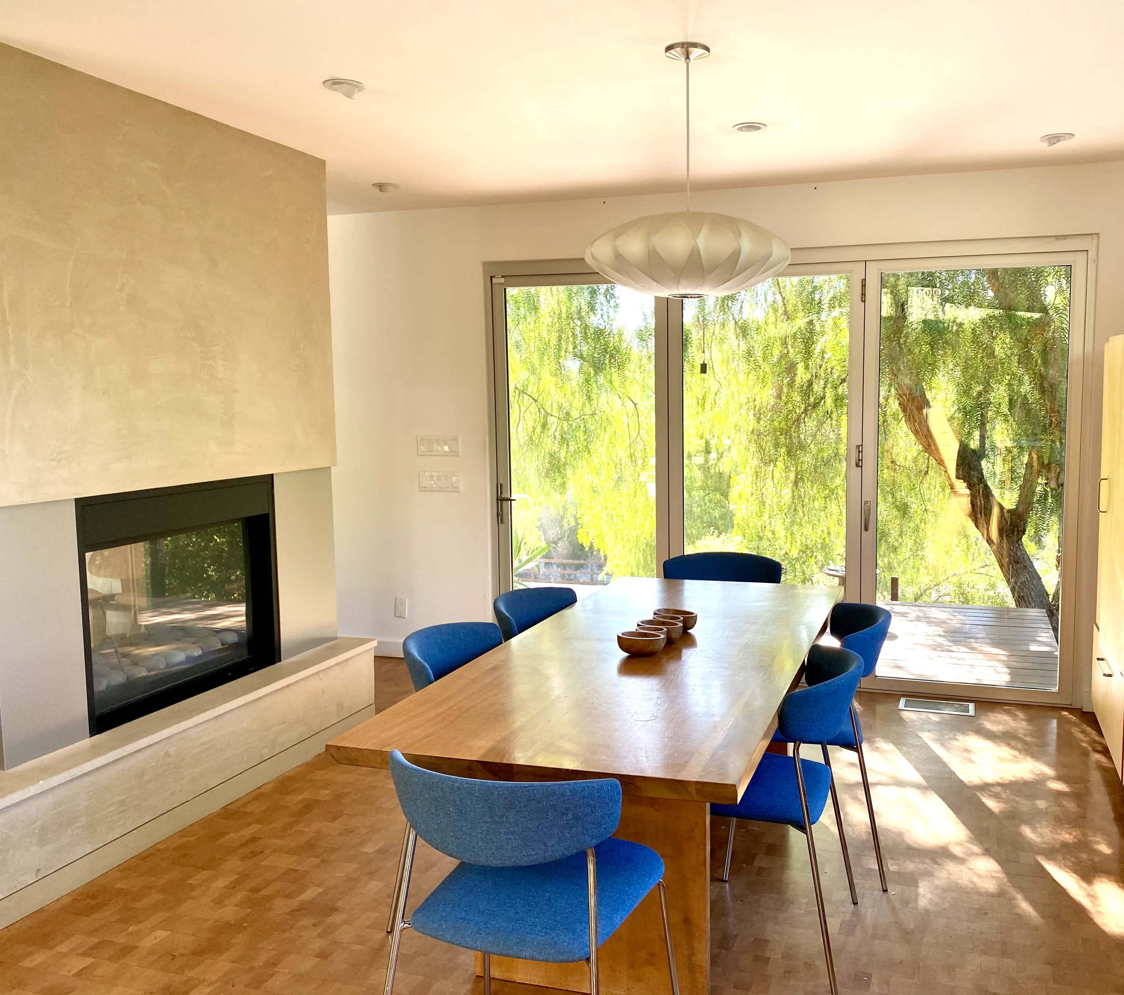 The image shows a modern dining room with a wooden table surrounded by blue chairs, a fireplace, and large windows revealing a tree outside.