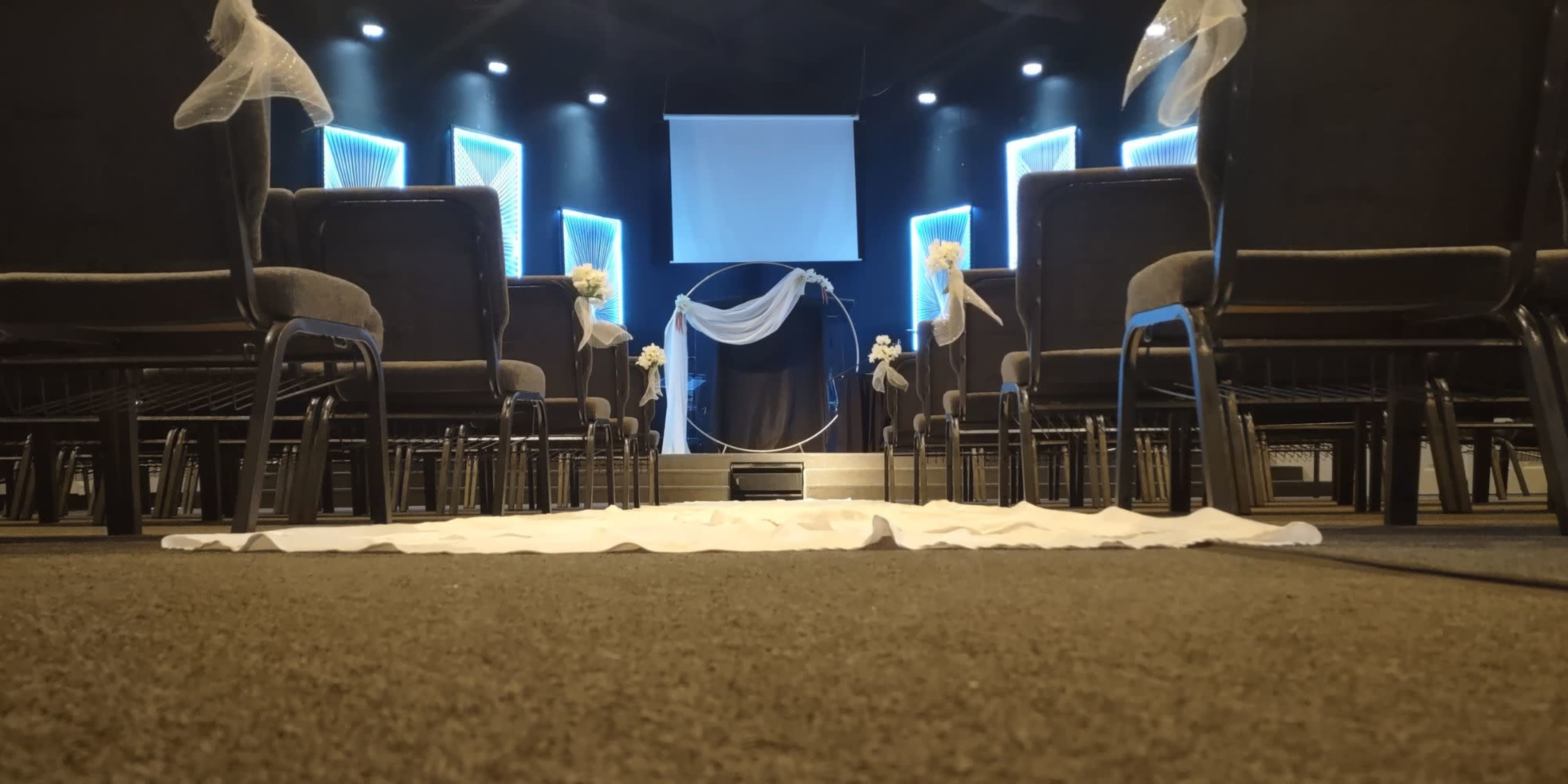 An indoor wedding venue with rows of chairs facing a decorated altar and a screen in the background.