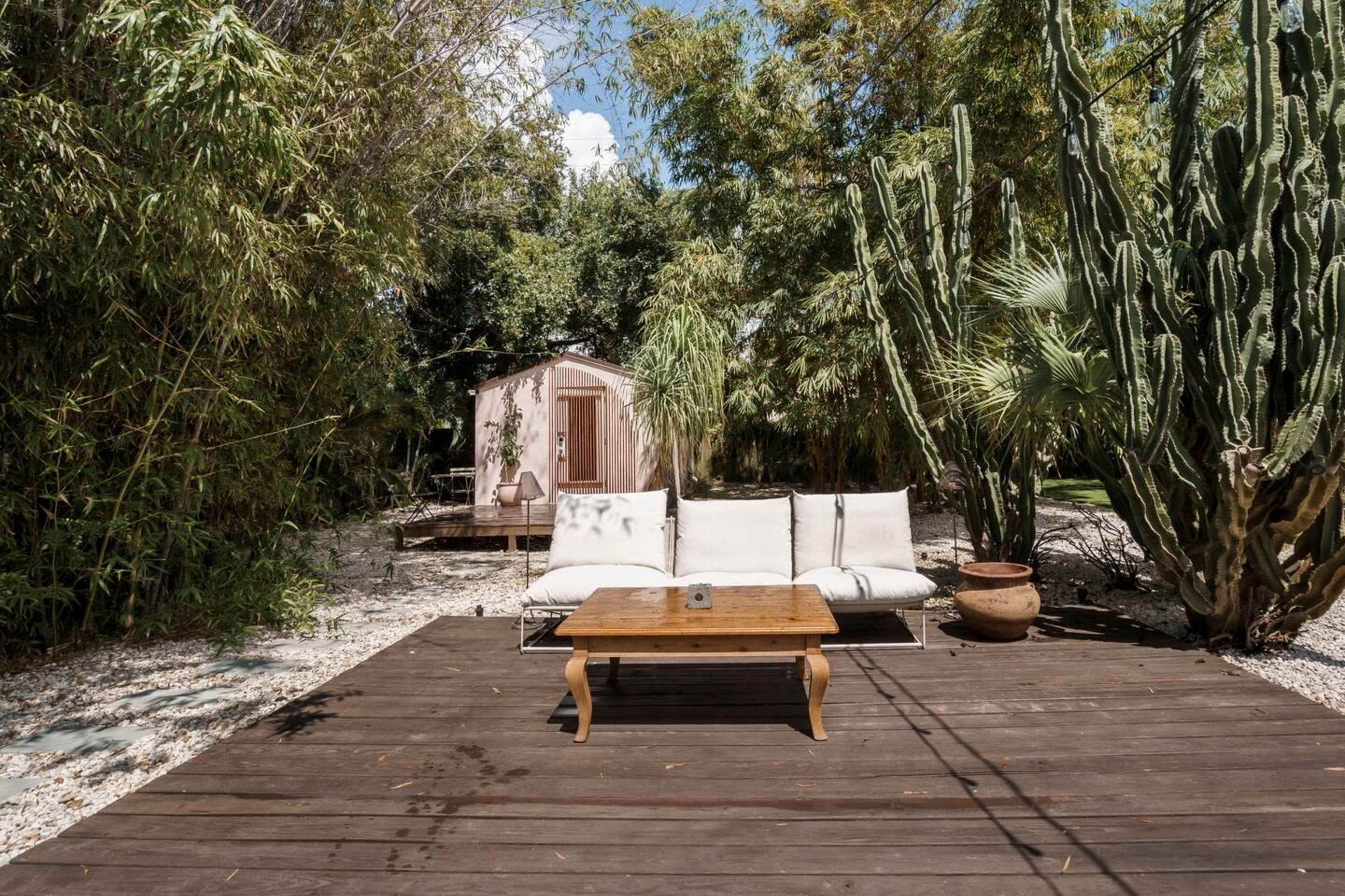 A wooden decking area with a light-colored sofa and a coffee table, surrounded by various plants and a small shed in the background.