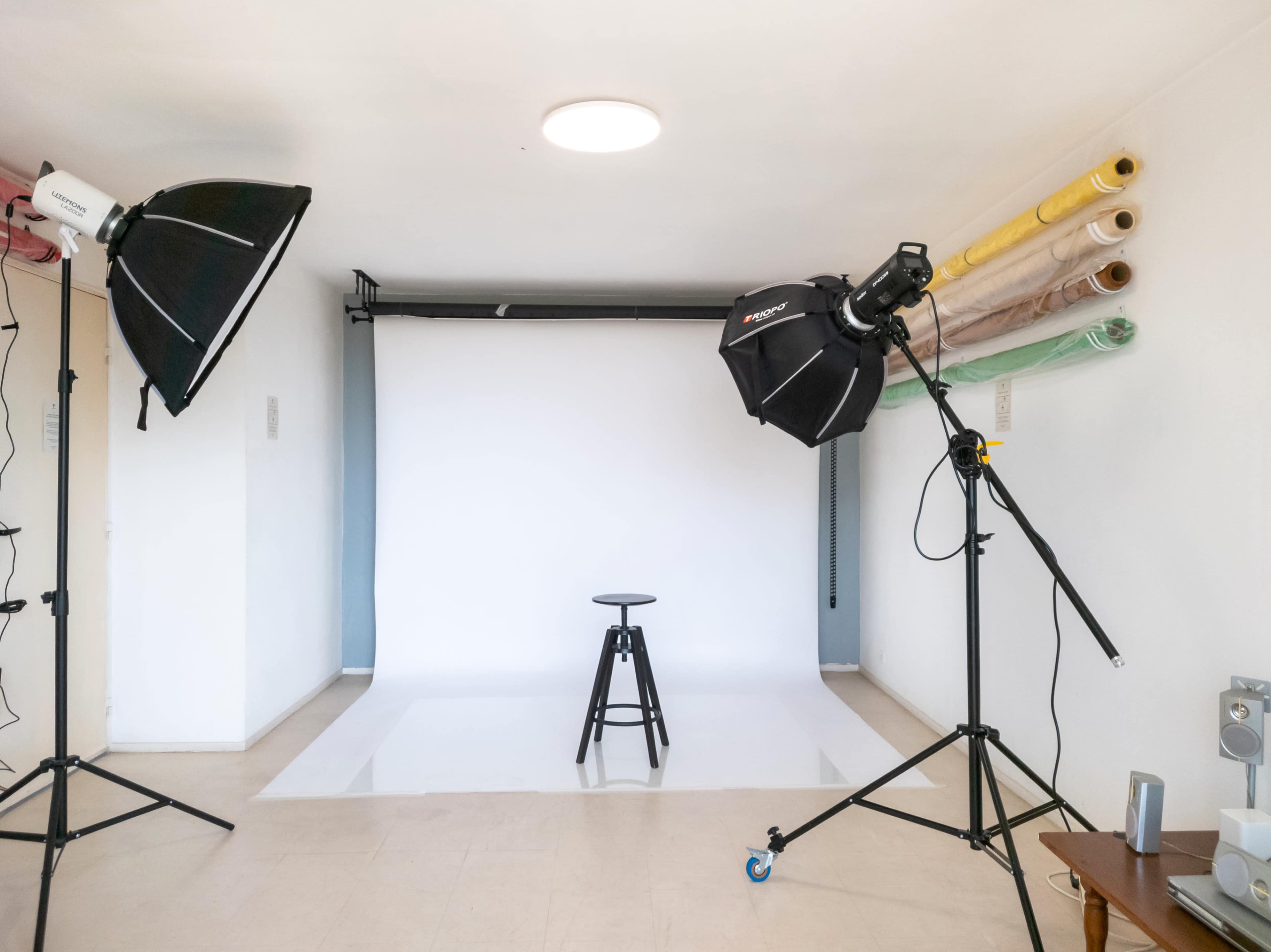 The image shows a photography studio with two lighting setups, a black stool in front of a white backdrop, and various rolls of paper in the background.