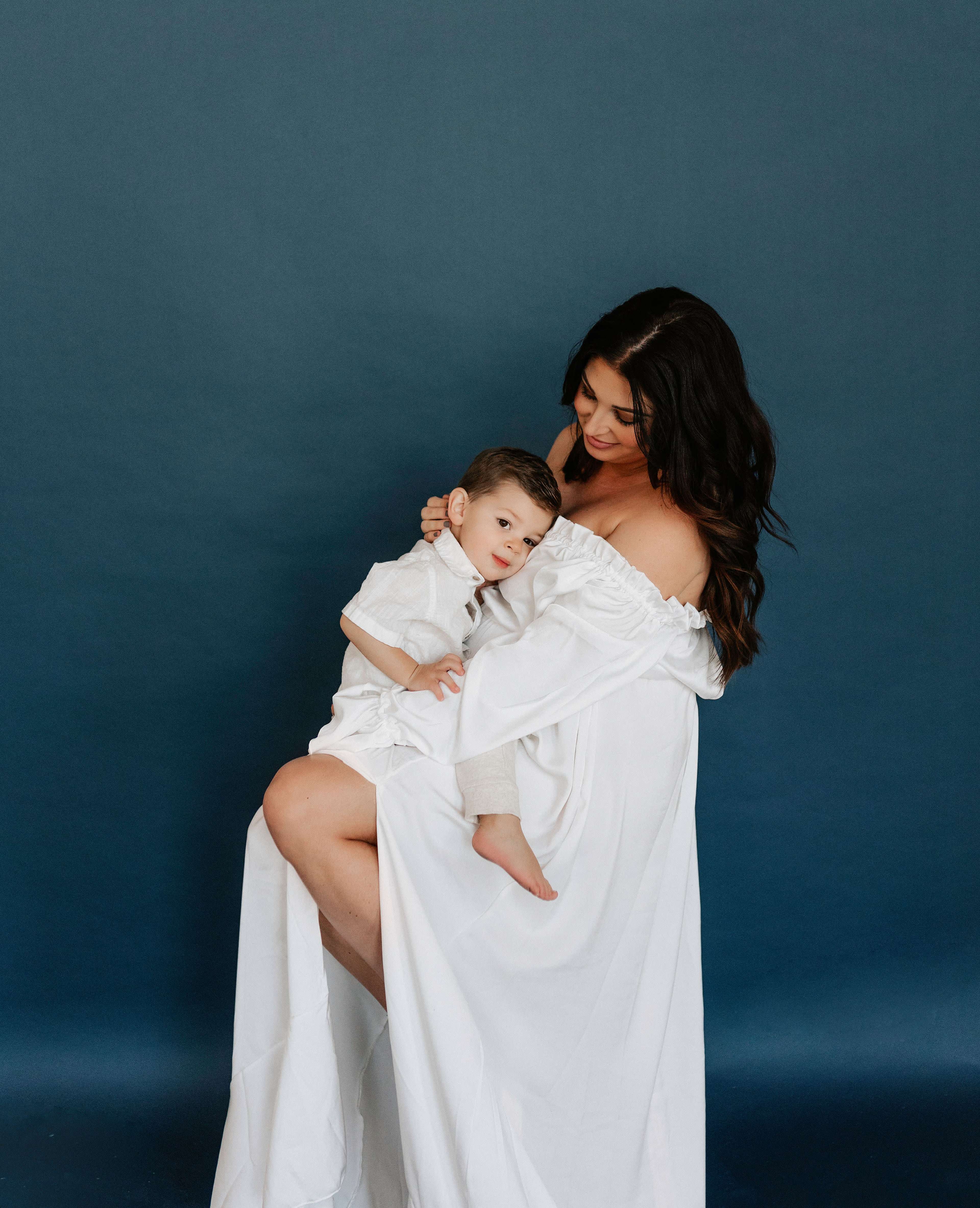 A woman holds a child in a white dress against a dark blue background.