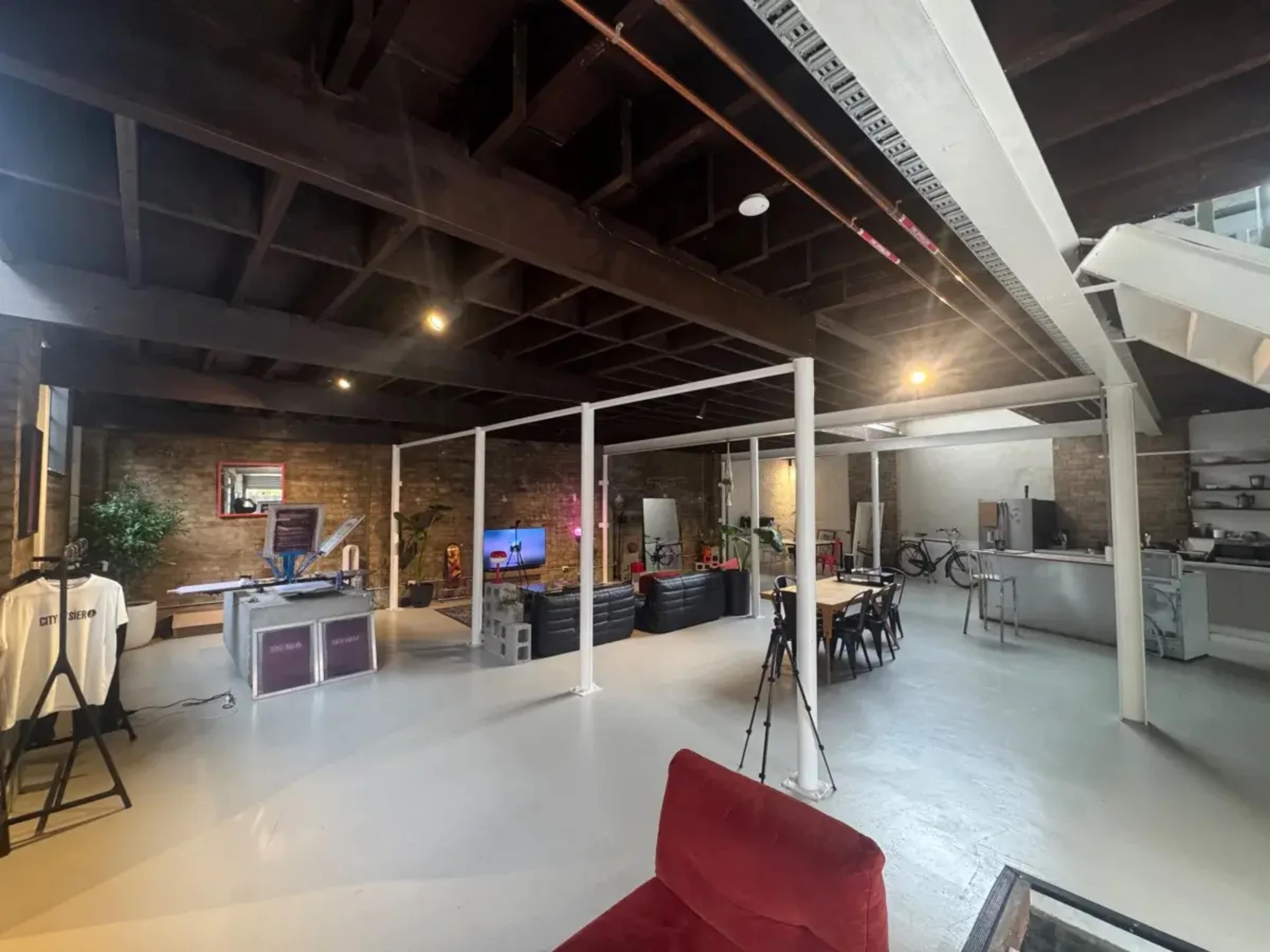 The image shows a spacious, modern open-concept office with exposed brick walls and various workstations, including a dining table and a kitchenette.