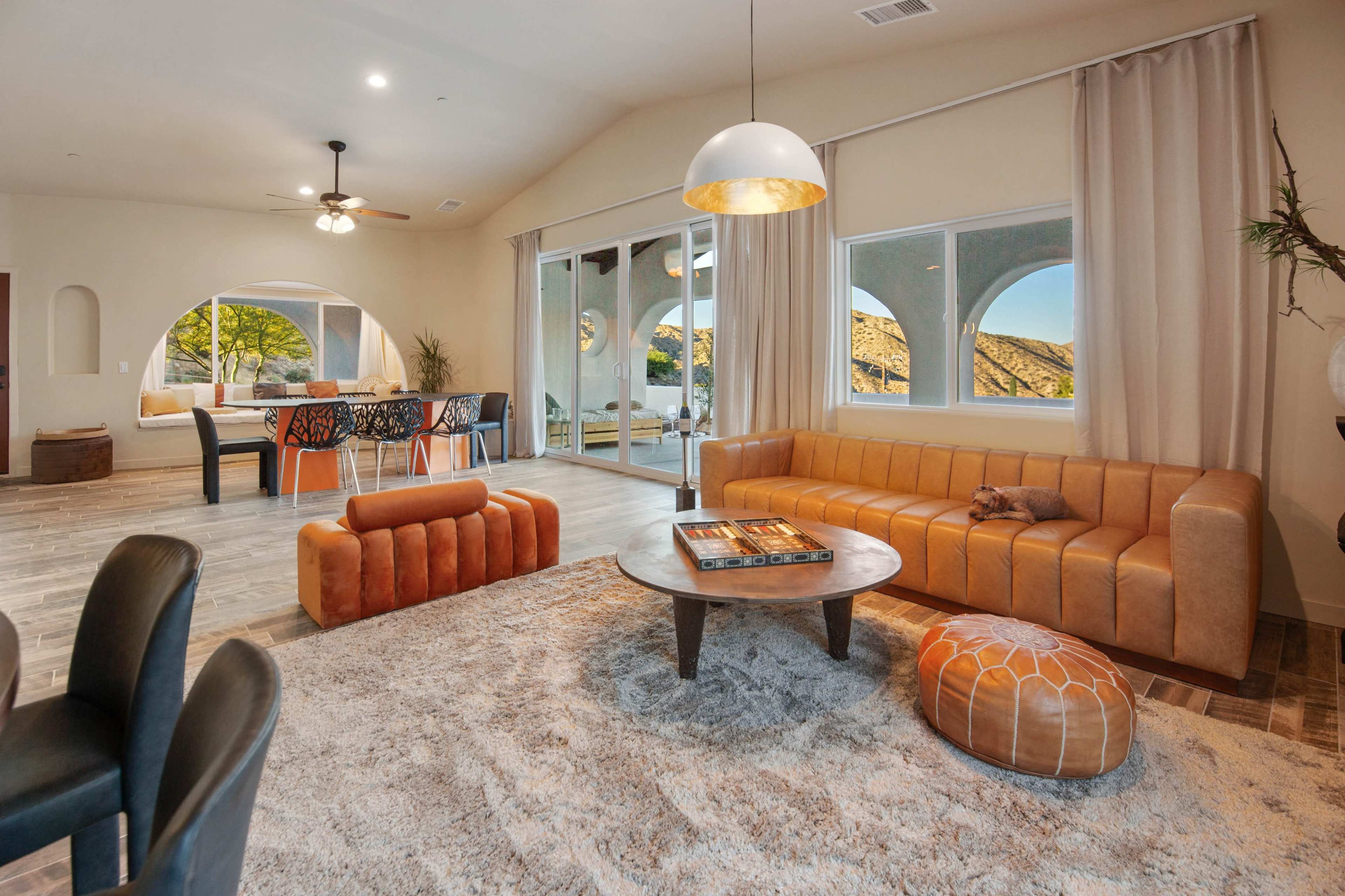 The image shows a modern living room with a large window overlooking a scenic view, featuring an orange sofa, a round coffee table, and wooden dining chairs in the background.