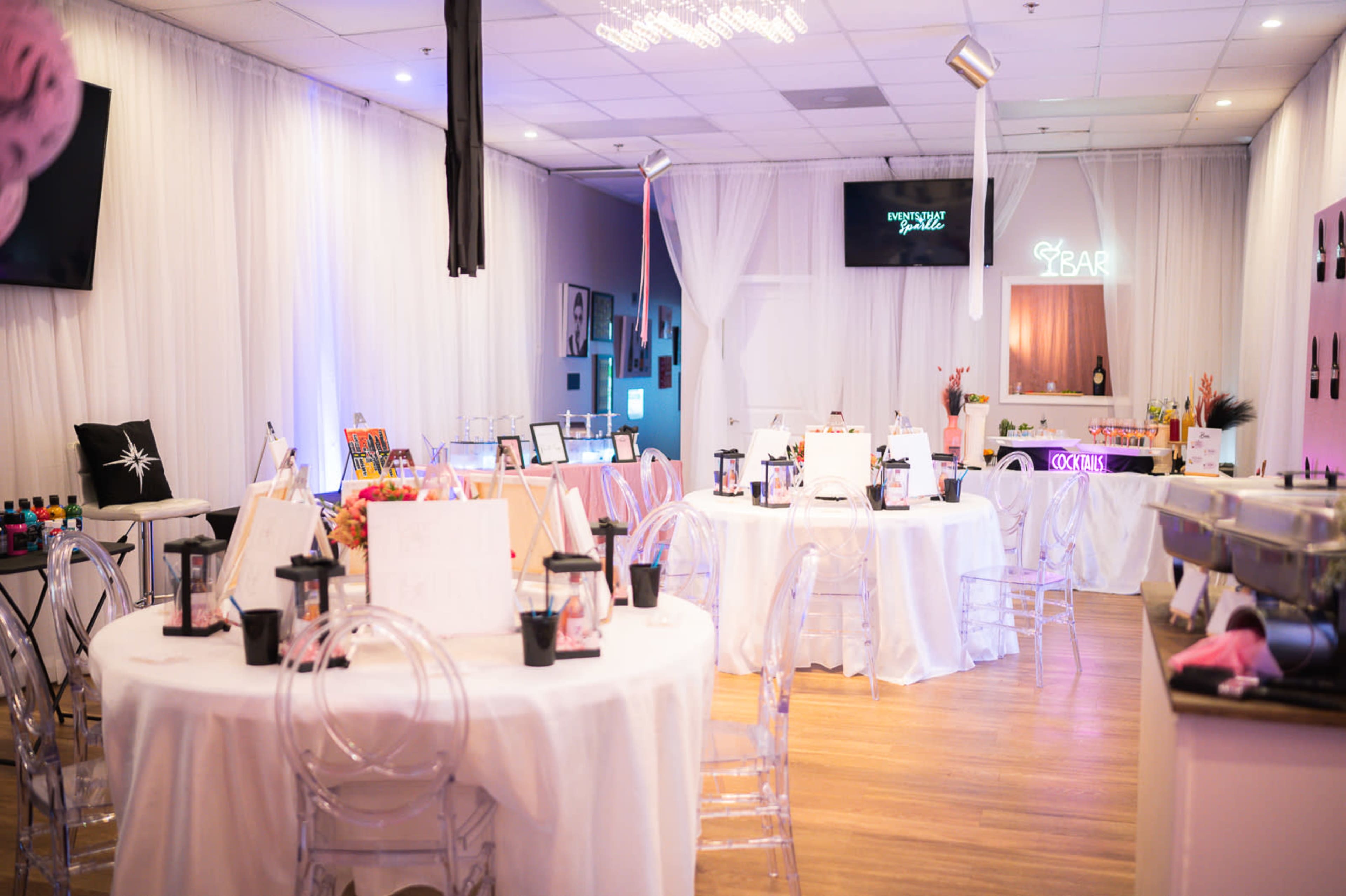 A decorated event space with several round tables set for a gathering, featuring white tablecloths, chairs, and a bar area in the background.