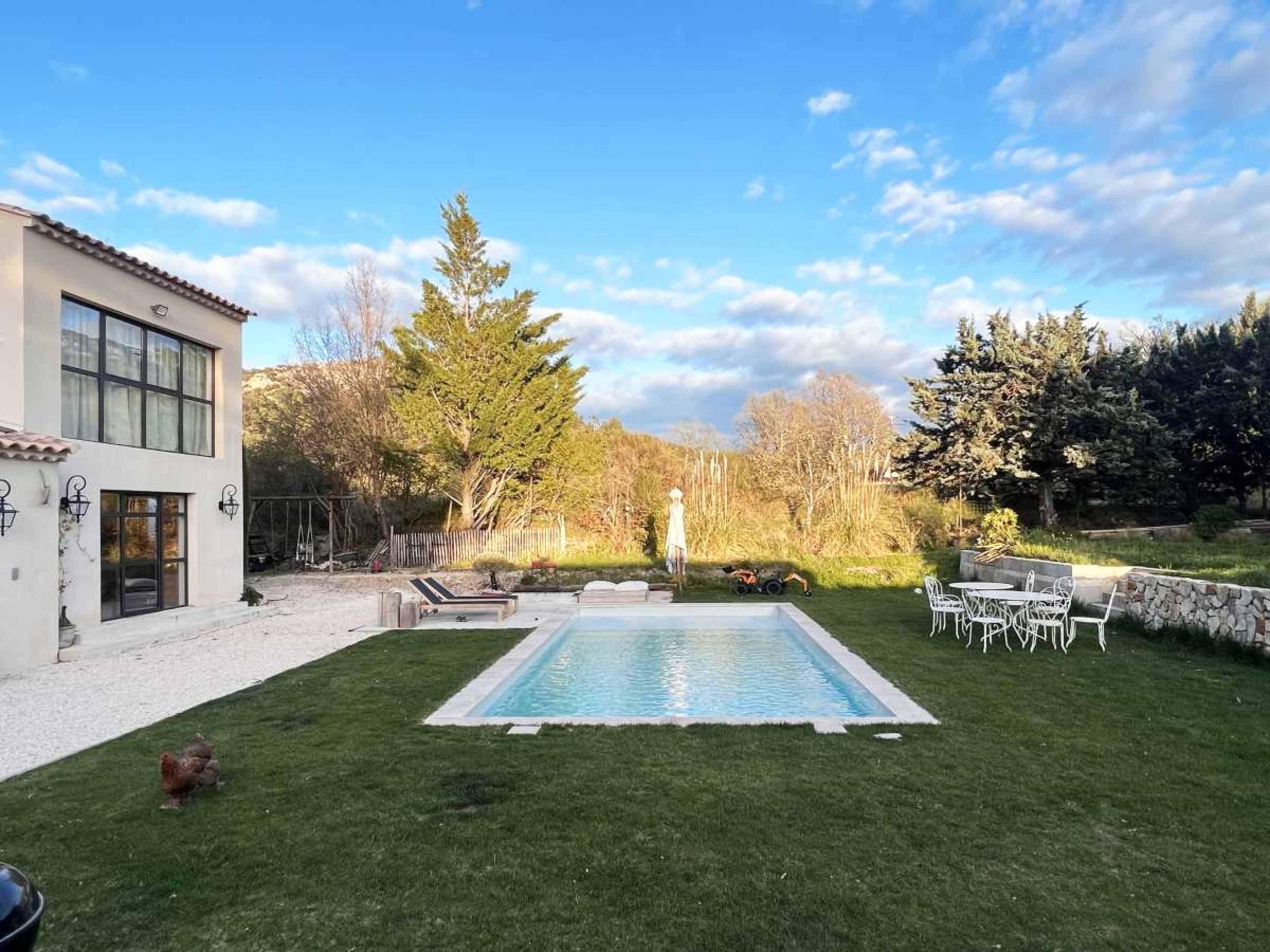 A well-maintained backyard features a swimming pool, a lawn with a few trees, and outdoor seating near a house.