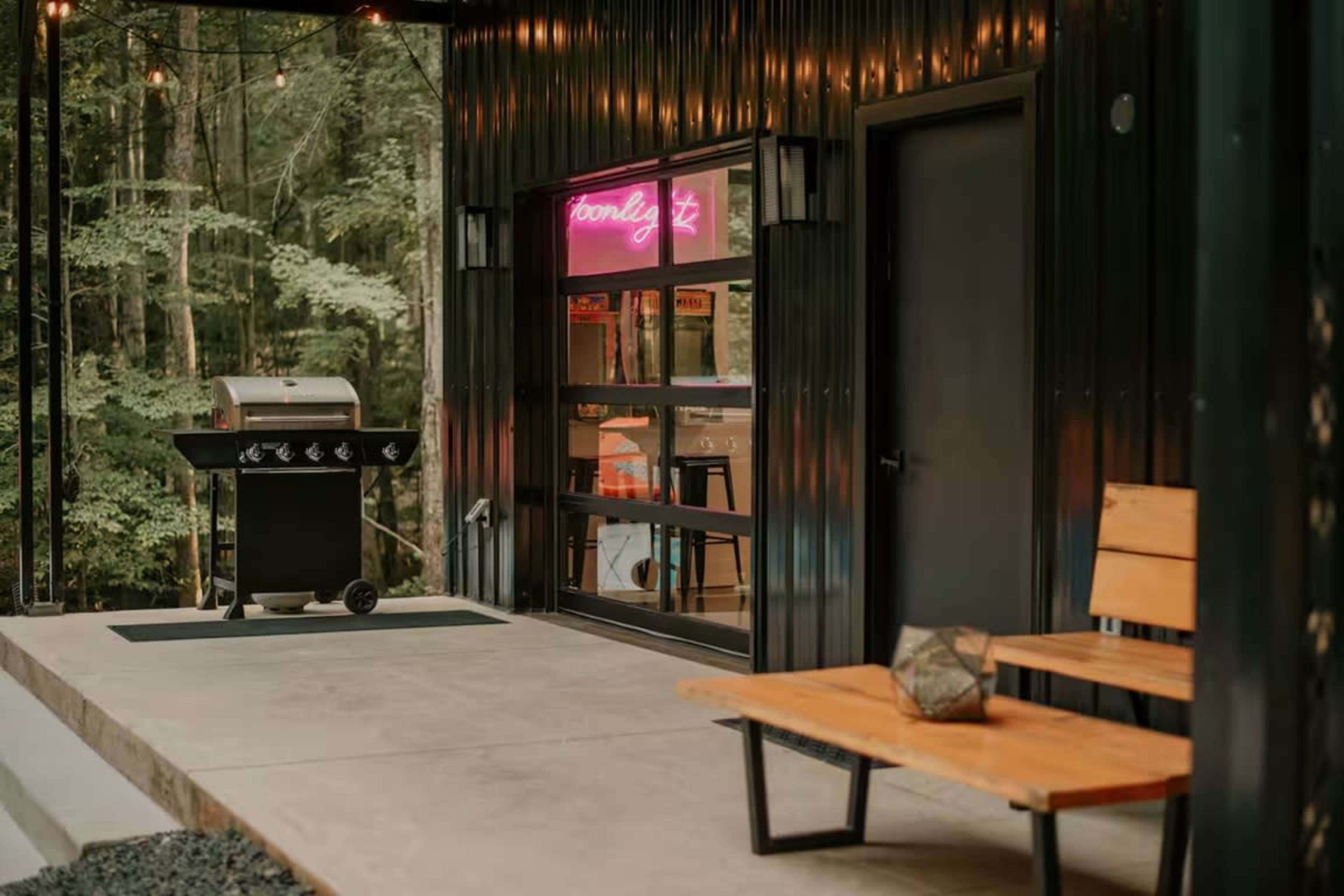 The image shows a modern outdoor area with a black exterior, featuring a bench, a gas grill, and a window illuminated by a neon sign.