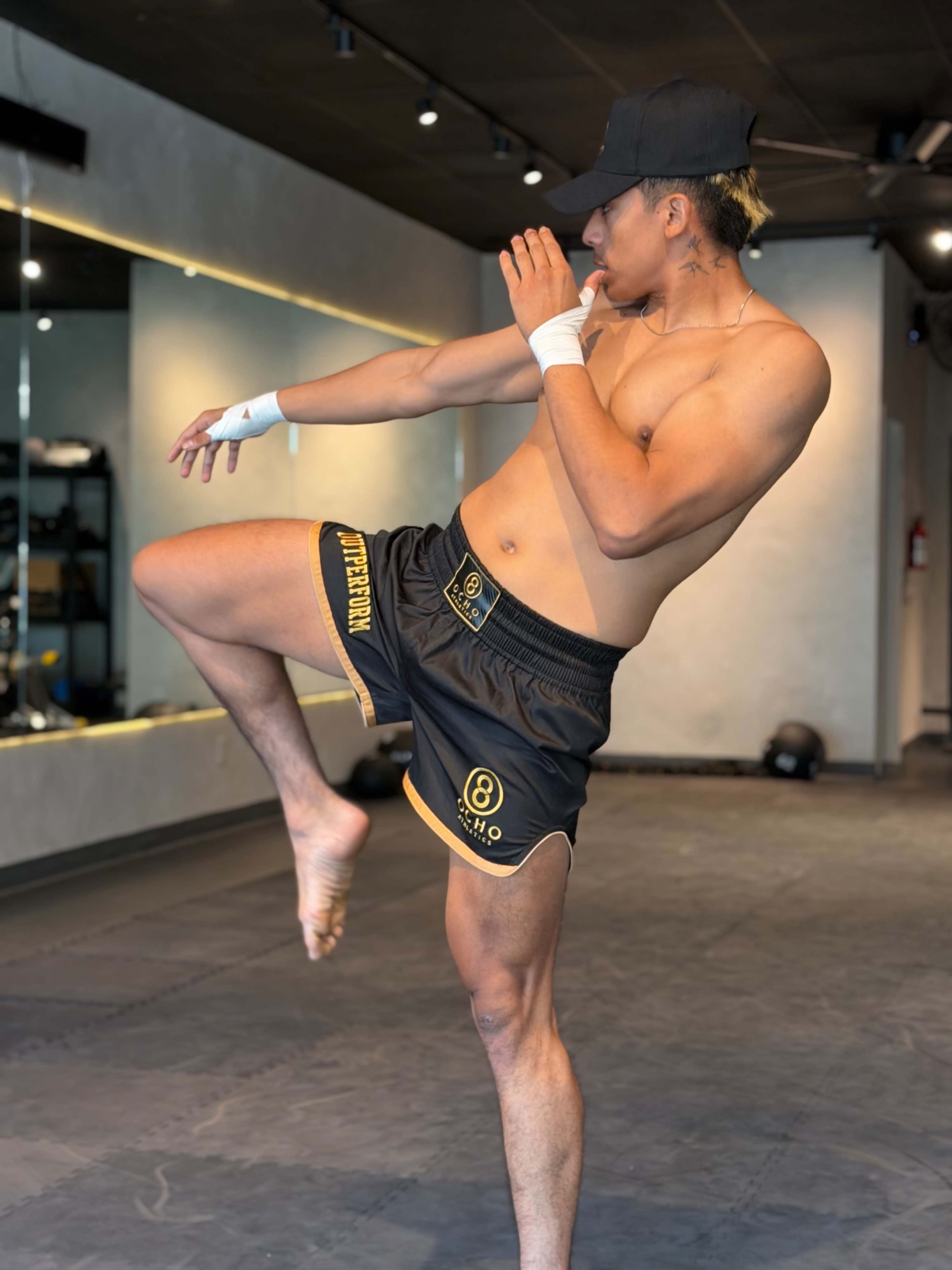 A shirtless man wearing black and yellow shorts performs a defensive stance in a gym setting.