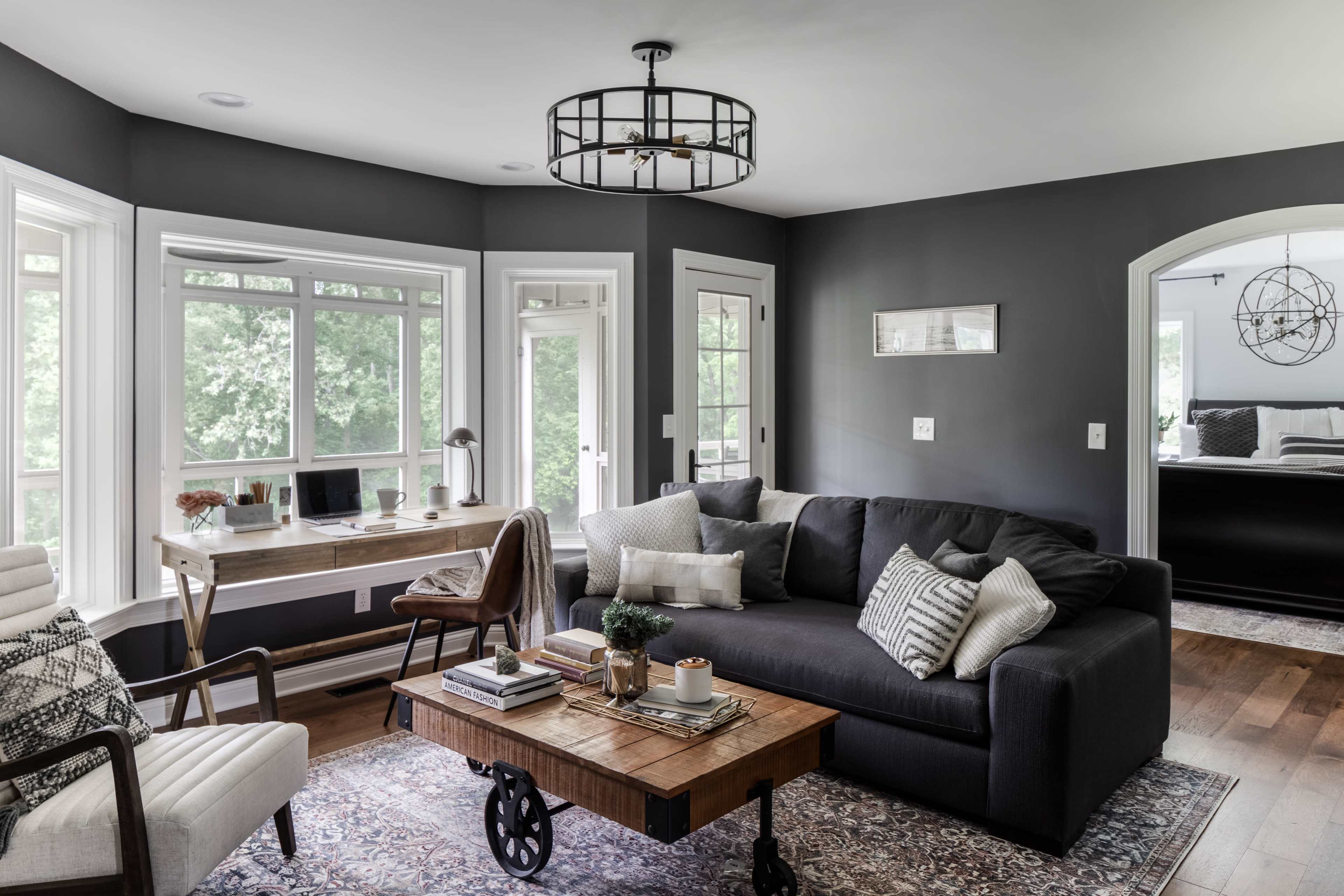 A modern, stylish living room featuring a gray sofa, a wooden coffee table on wheels, and a desk by a large window.