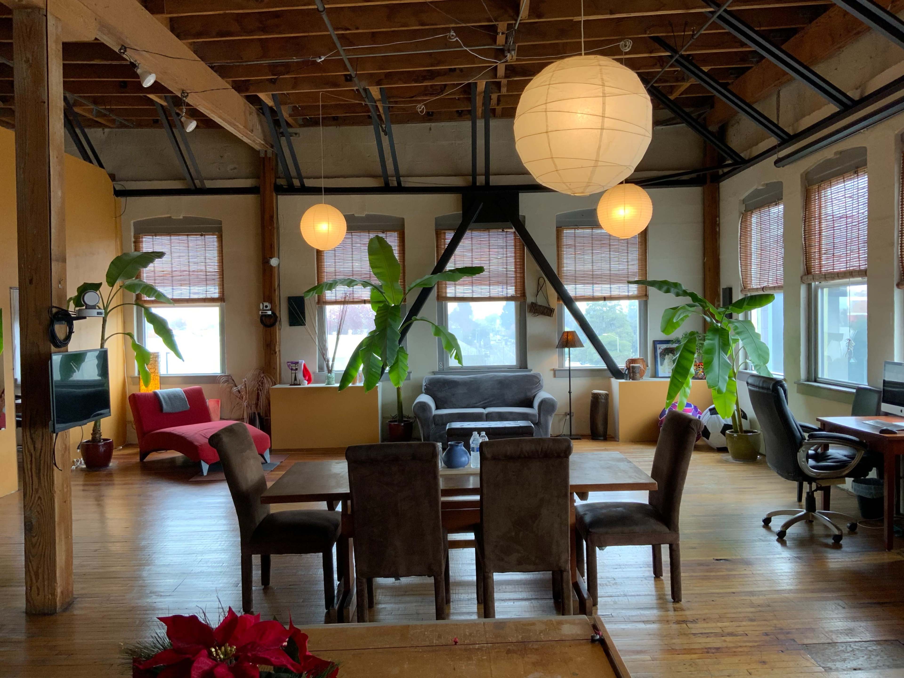 A spacious, open-concept living area furnished with a dining table, chairs, a grey couch, and several indoor plants, illuminated by large paper lanterns.