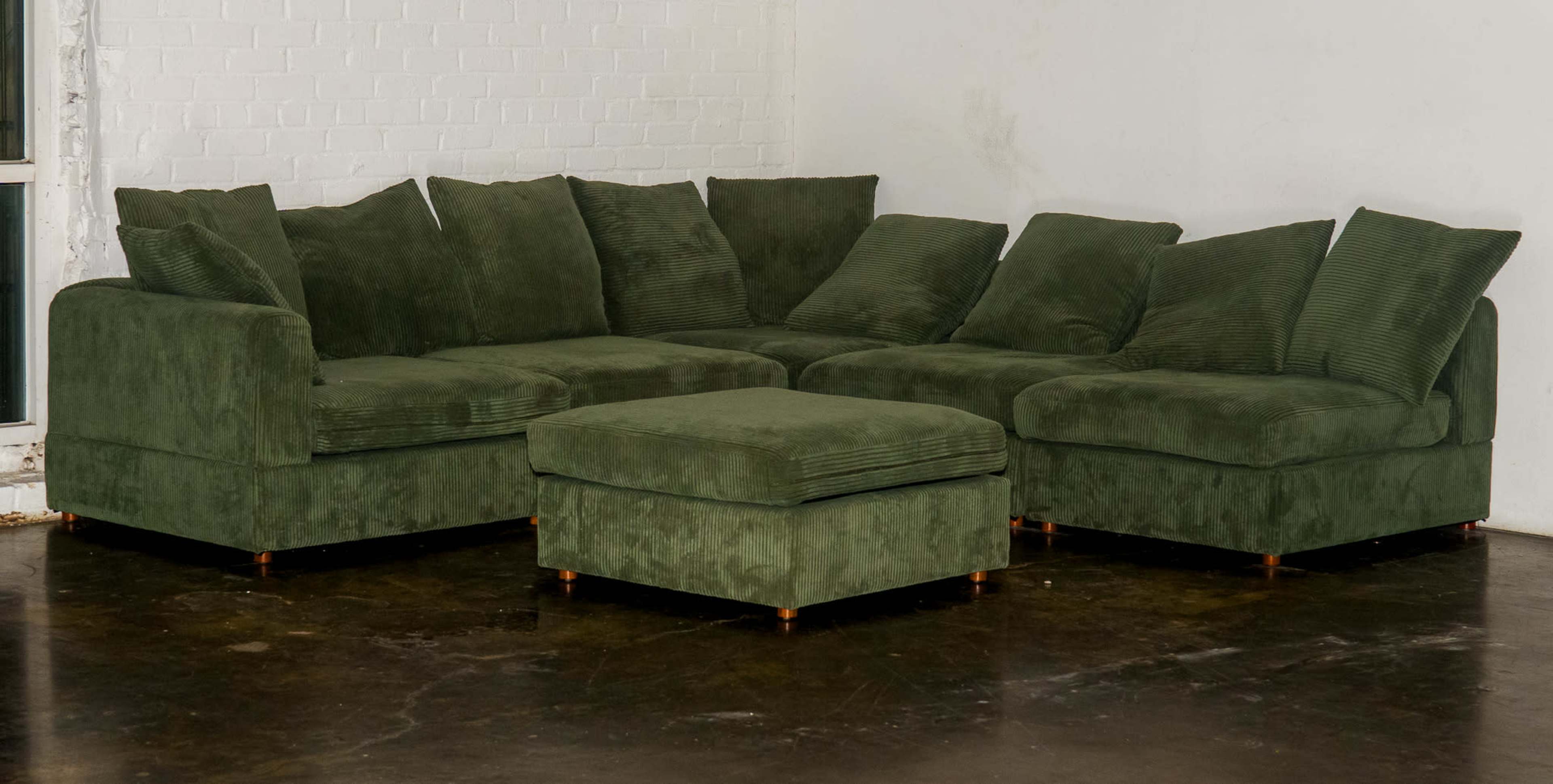 The image shows a large, green sectional sofa with several cushions and a matching ottoman arranged in a spacious interior setting.
