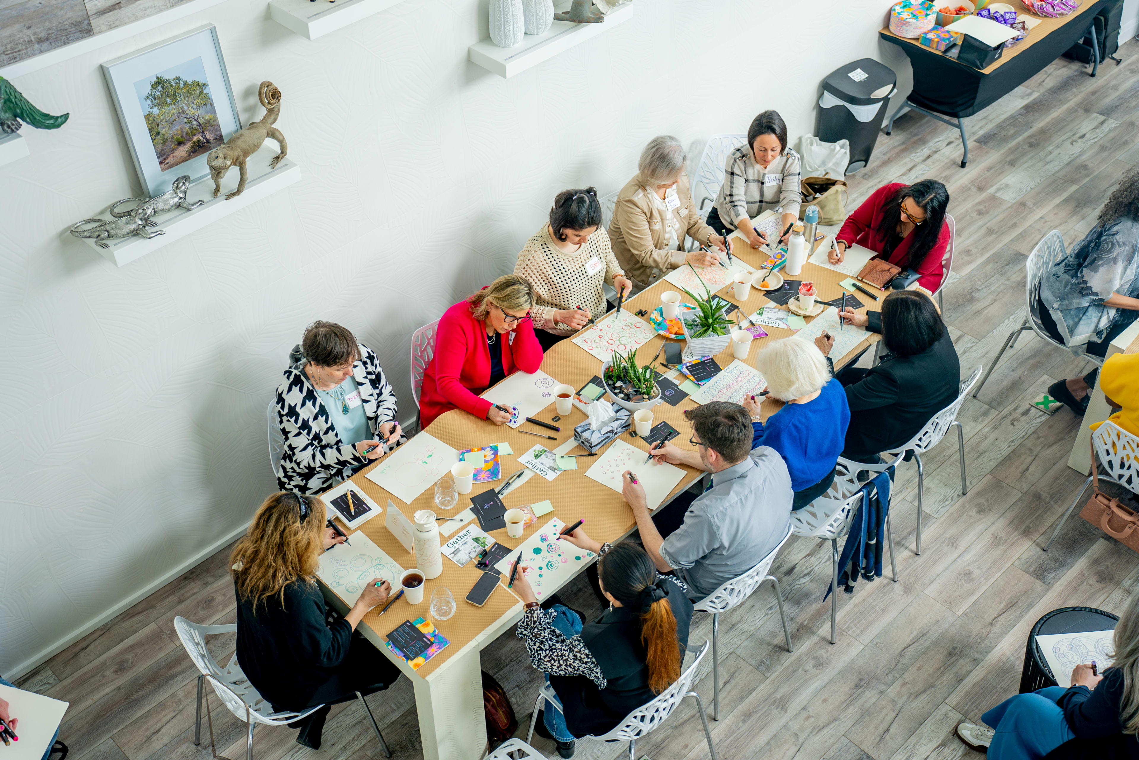 A group of people are seated around a long table engaged in an arts and crafts activity in a well-lit room.