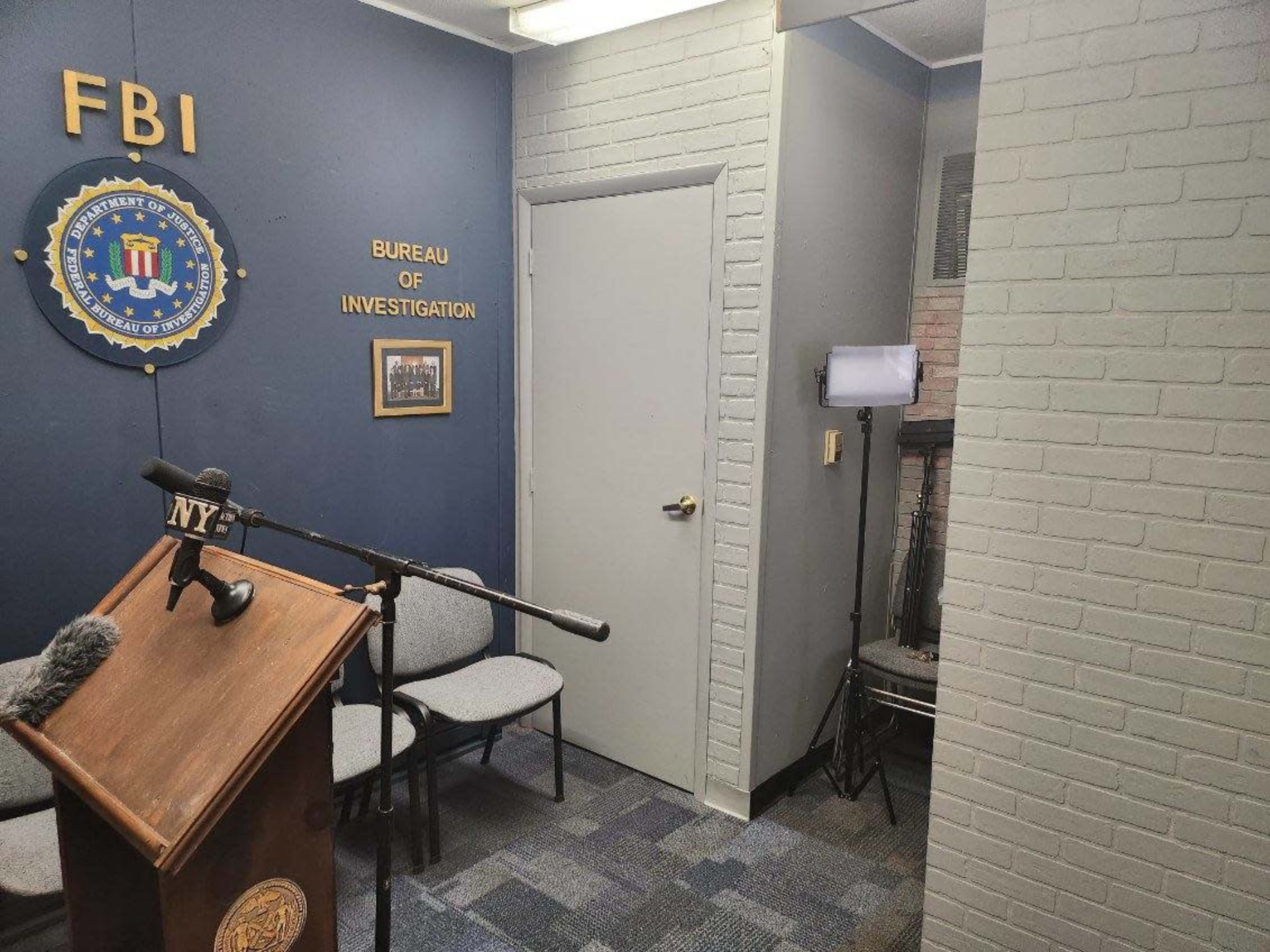 A small room with a podium featuring a microphone, chairs arranged in front of it, and a blue wall displaying an FBI seal.