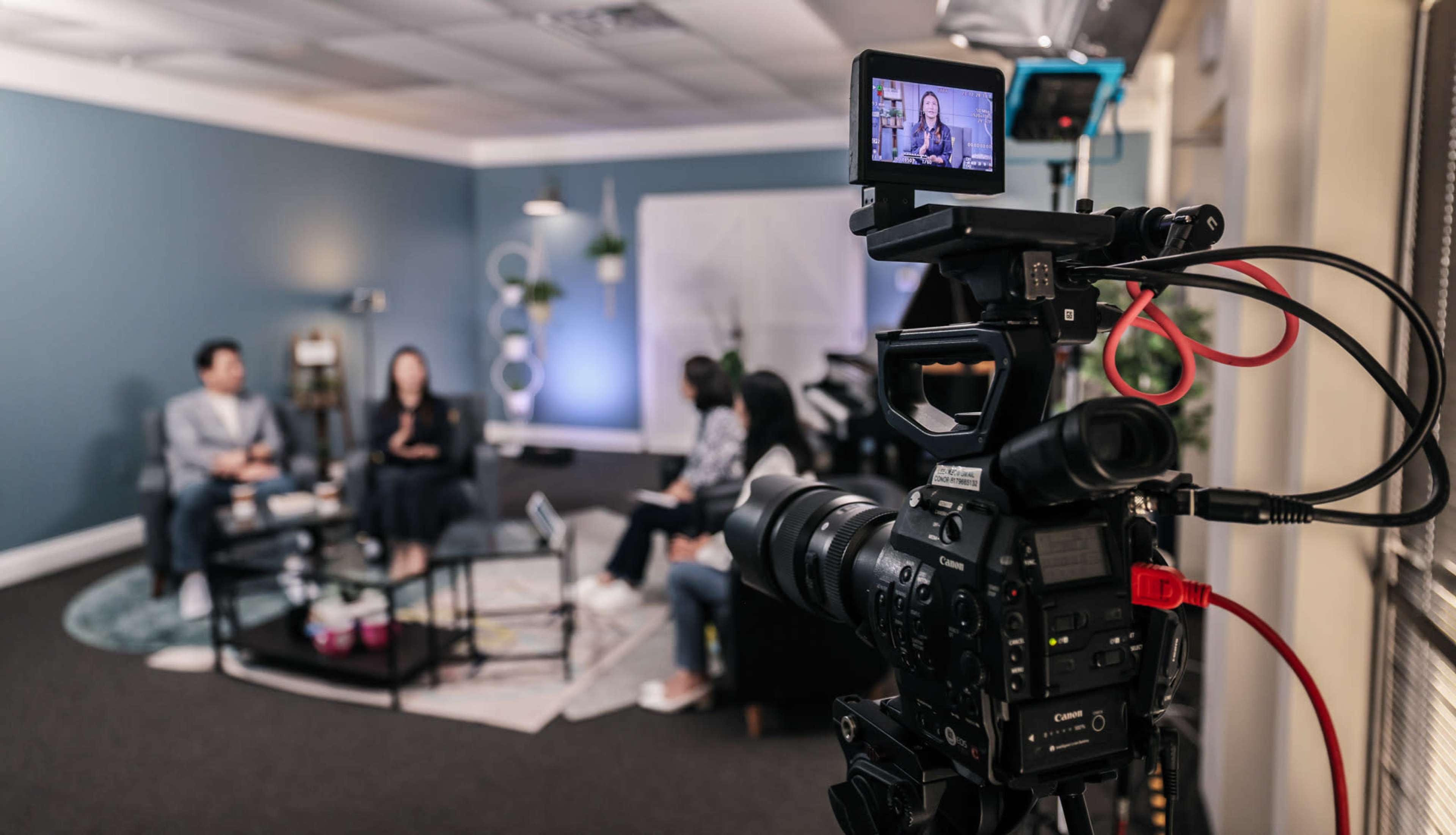 A camera captures a discussion between two people seated at a table while others observe in a well-lit room.