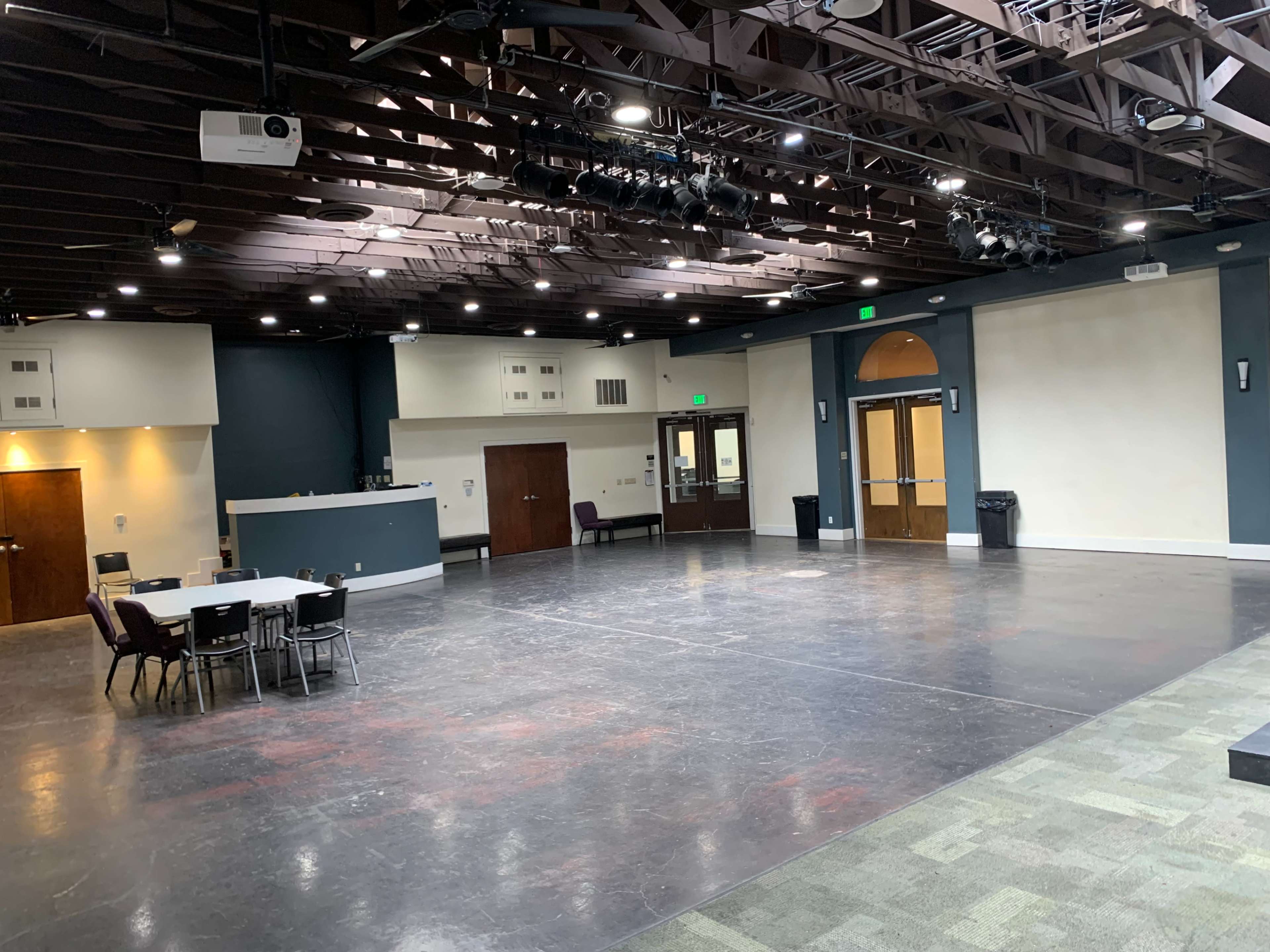 The image shows a large, empty multipurpose room with a dark floor, overhead lighting, and a small stage area, featuring tables and chairs arranged on one side.