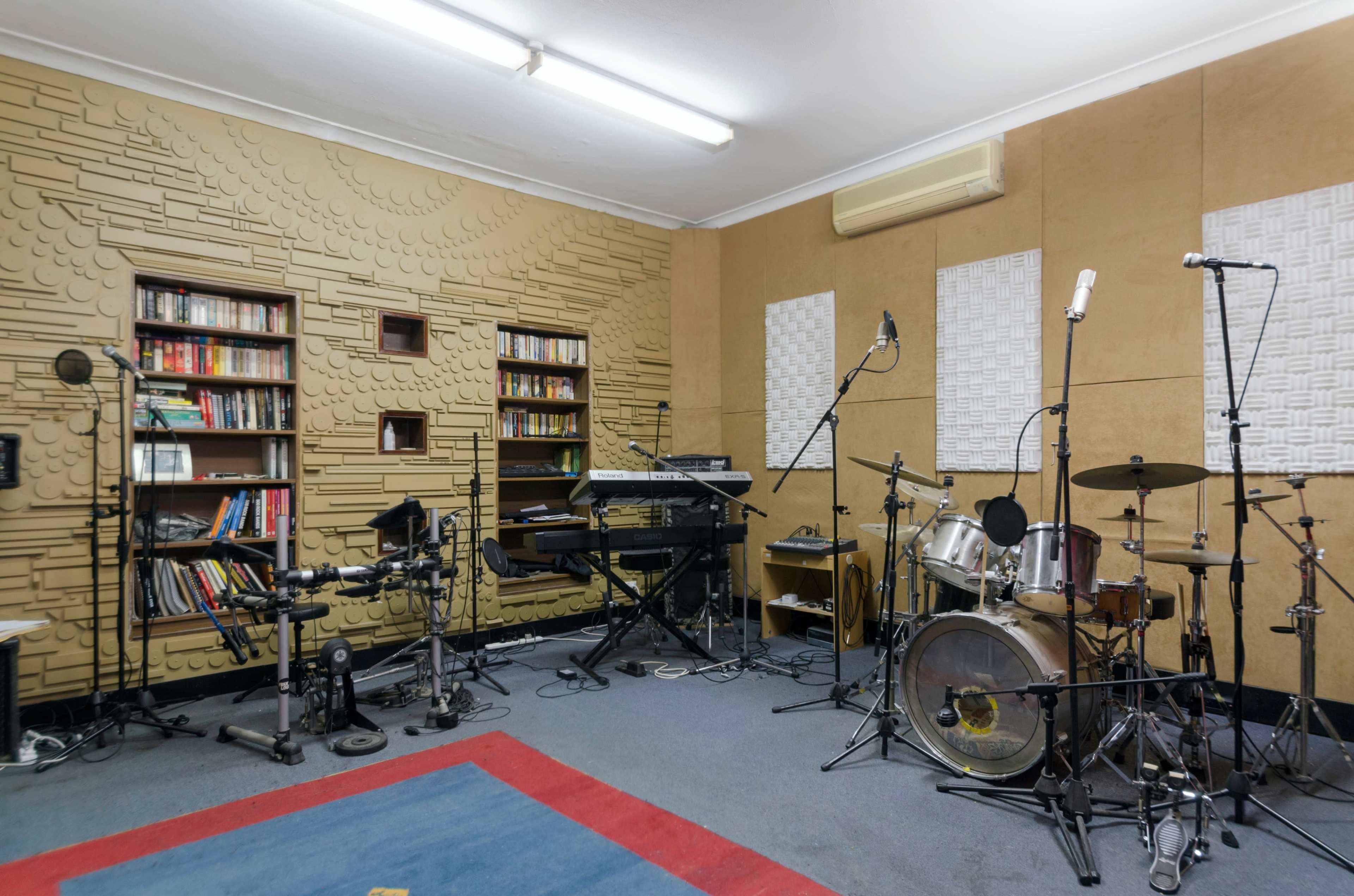 The image shows a music recording studio with a variety of instruments, including a drum set, keyboard, and electronic drum pads, alongside soundproofing panels on the walls.