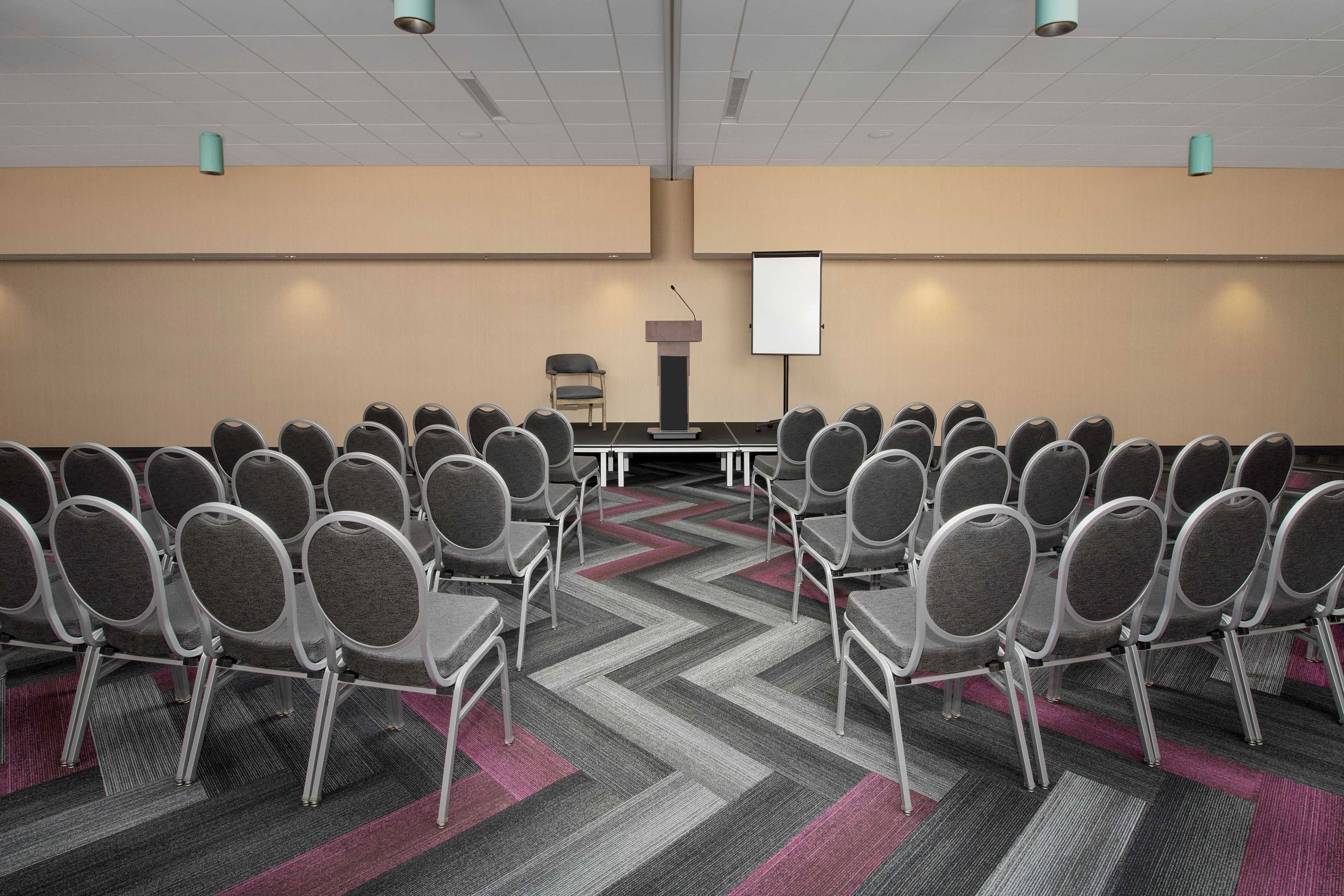 A room is set up for a presentation with rows of empty circular chairs facing a podium and a blank presentation screen.