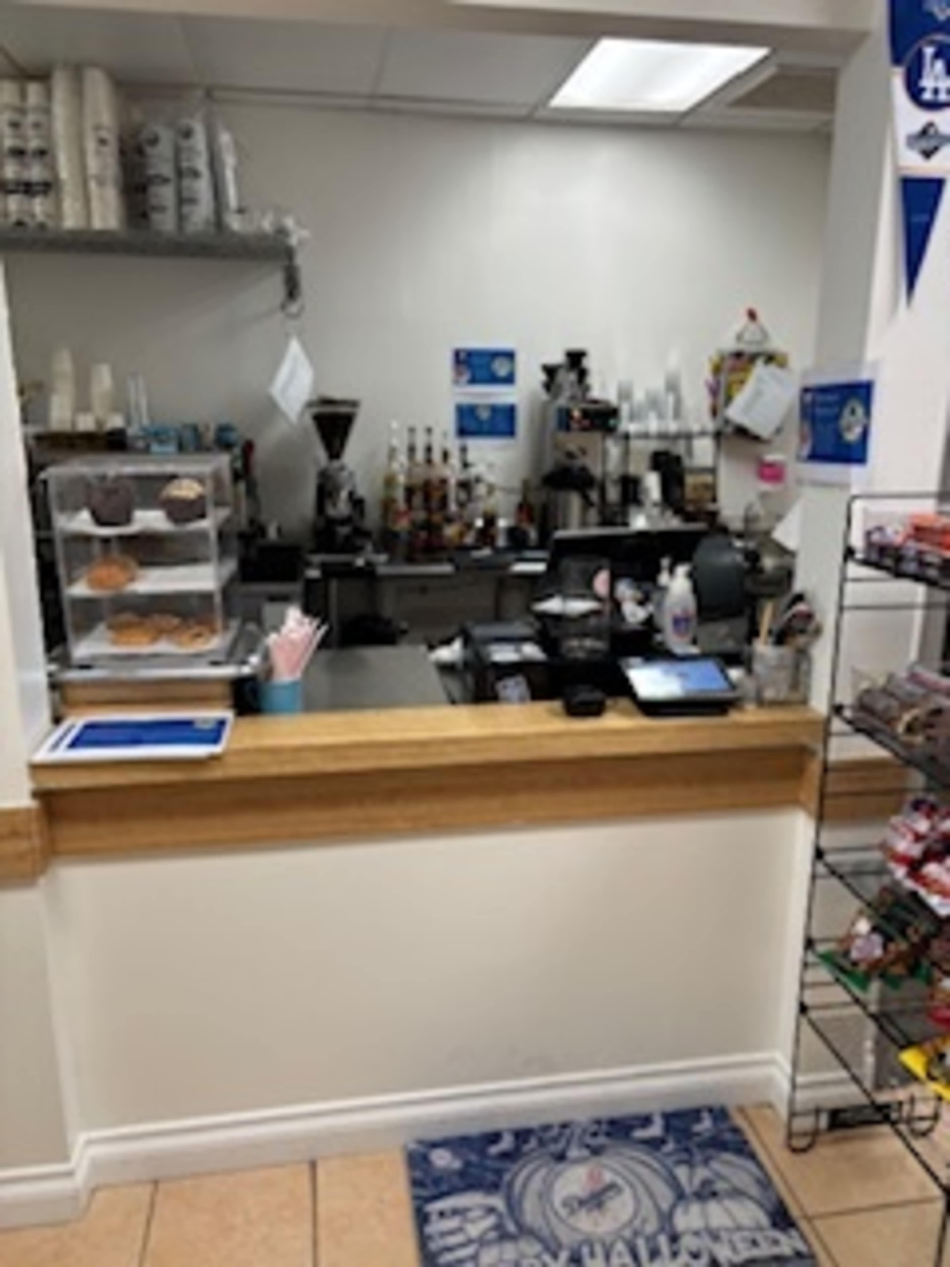 The image shows a small coffee shop counter with a display case for pastries, a coffee maker, and a shelf stocked with cups and supplies.