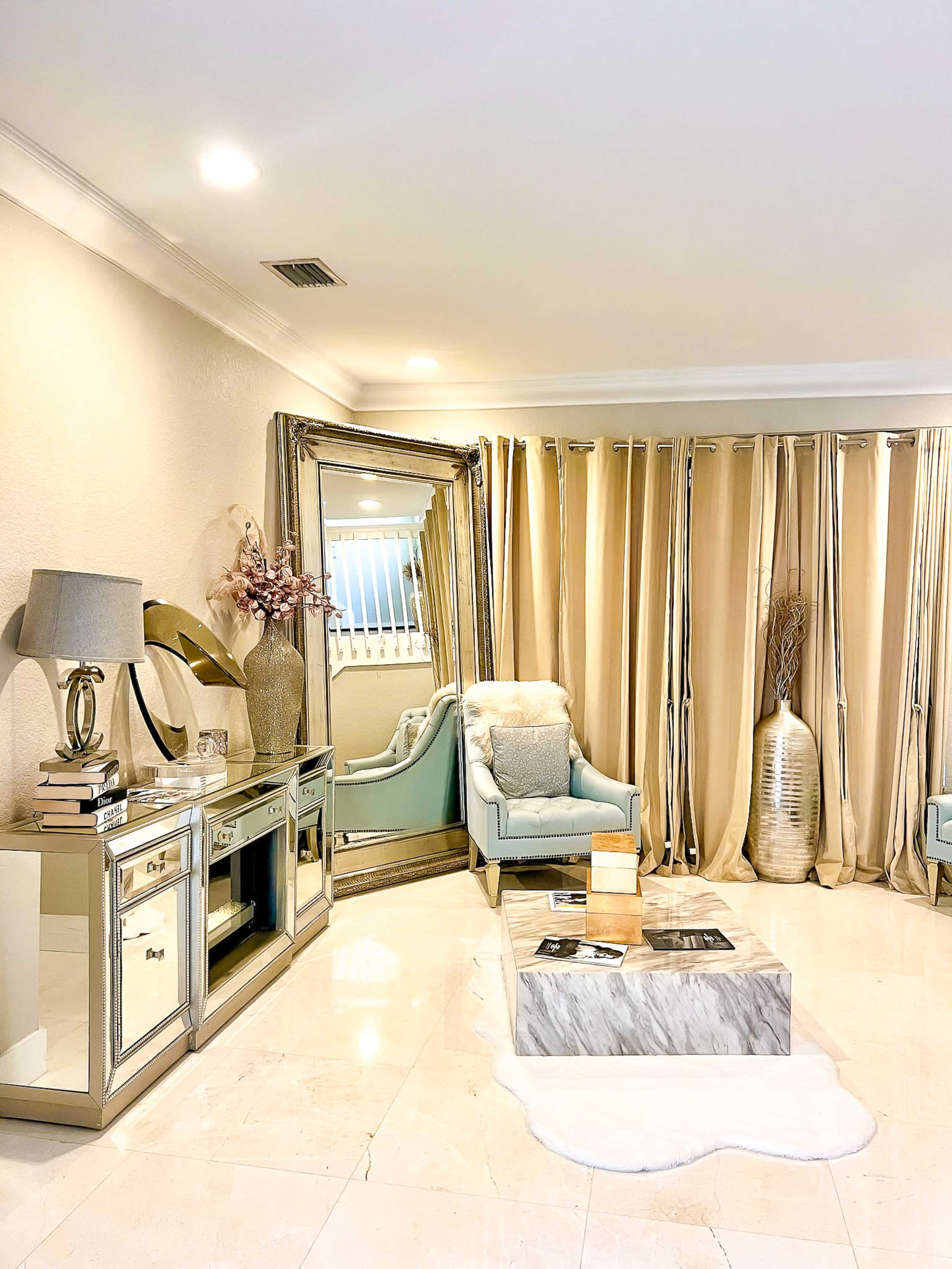 The image shows a stylish living room featuring a light-colored marble coffee table, a mirrored cabinet, a blue accent chair, and beige curtains.