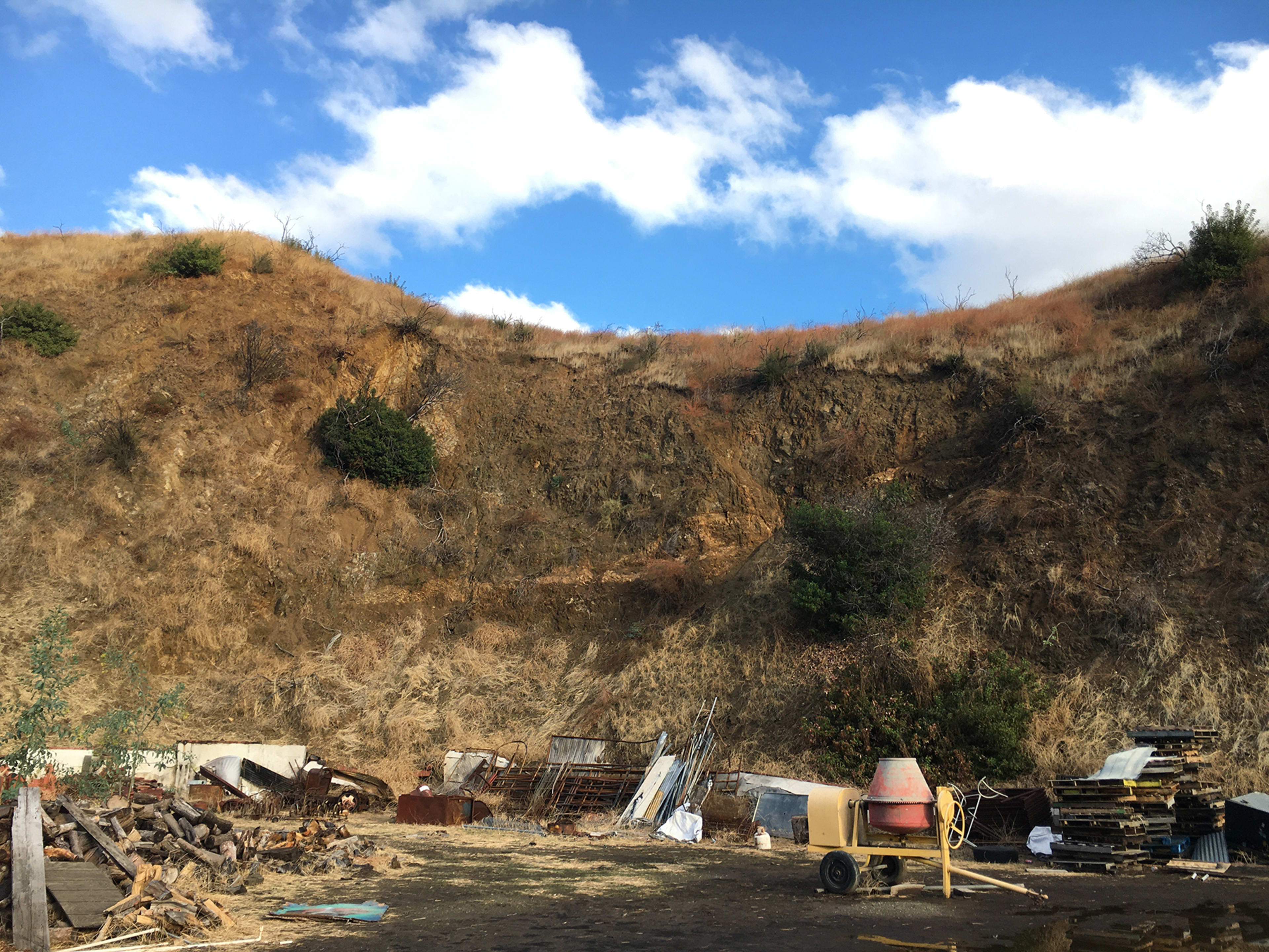 The image shows a hillside covered with dry grass and sparse vegetation, with scattered debris and structures visible at the base.