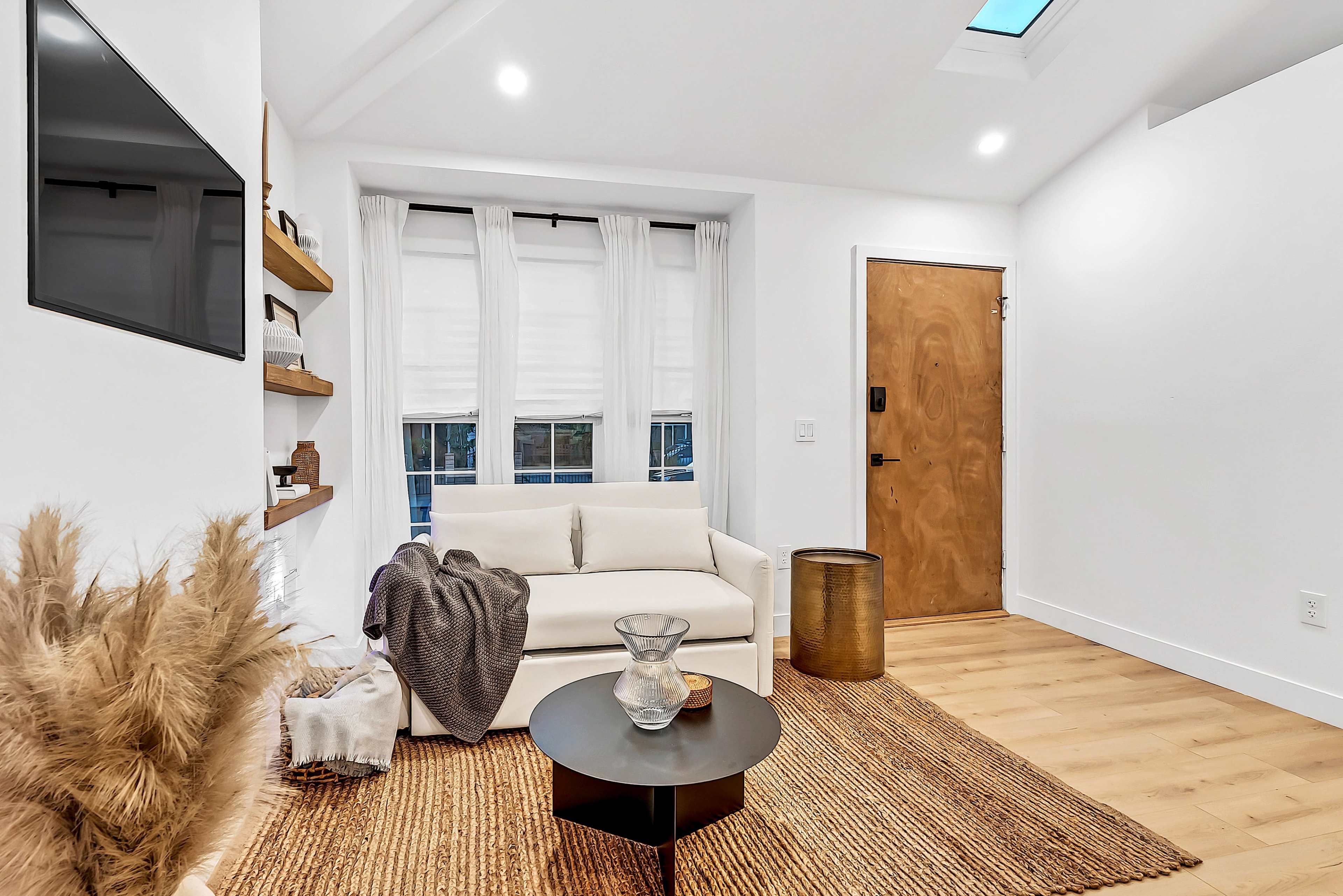The image shows a modern living room featuring a white sofa, a round black coffee table, a textured area rug, and a wooden door with a glass panel.