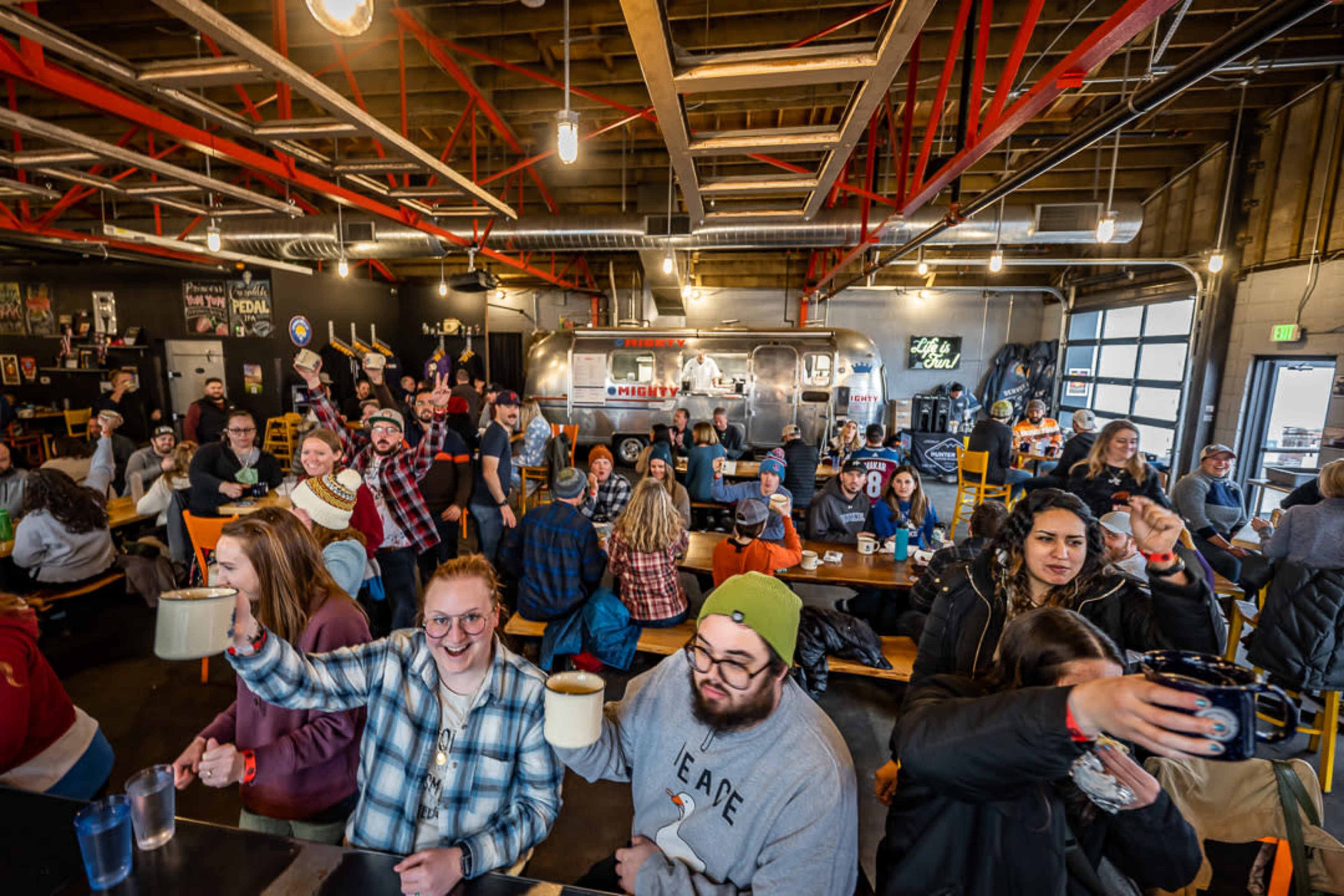 A lively crowd sits at long tables in a bustling indoor market, raising mugs and smiling at the camera.