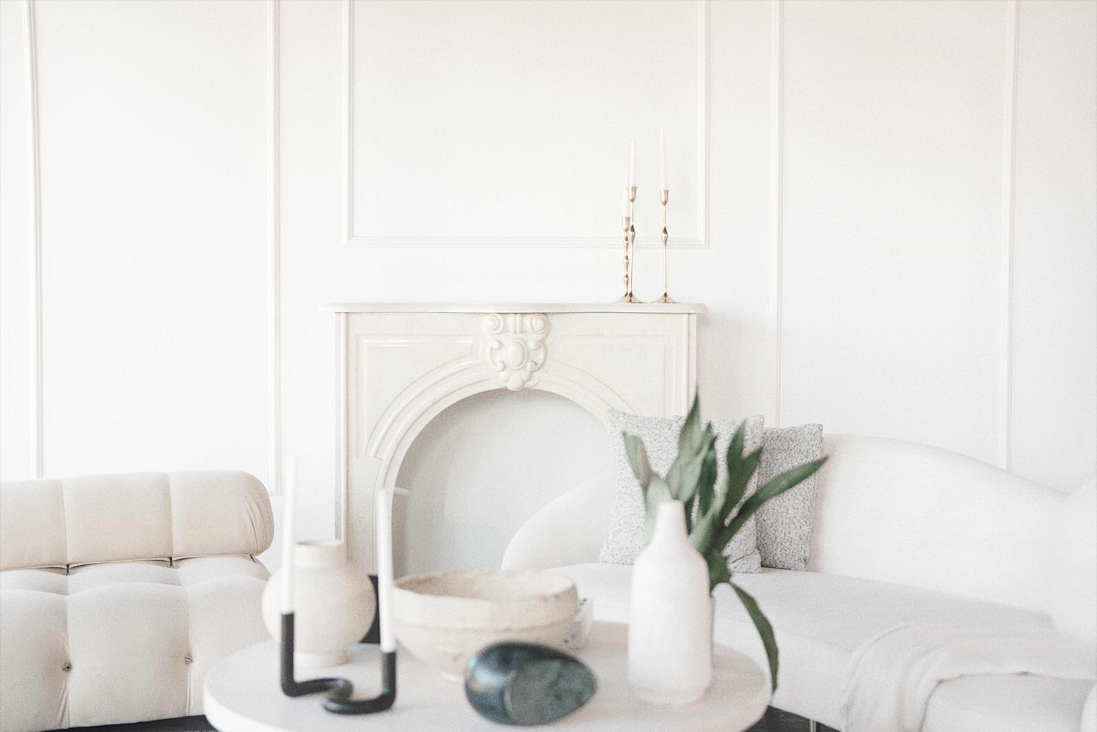 The image shows a bright, minimalist living space featuring a white sofa, a light-colored tufted chair, and a decorative fireplace, with a round table displaying various decor items.