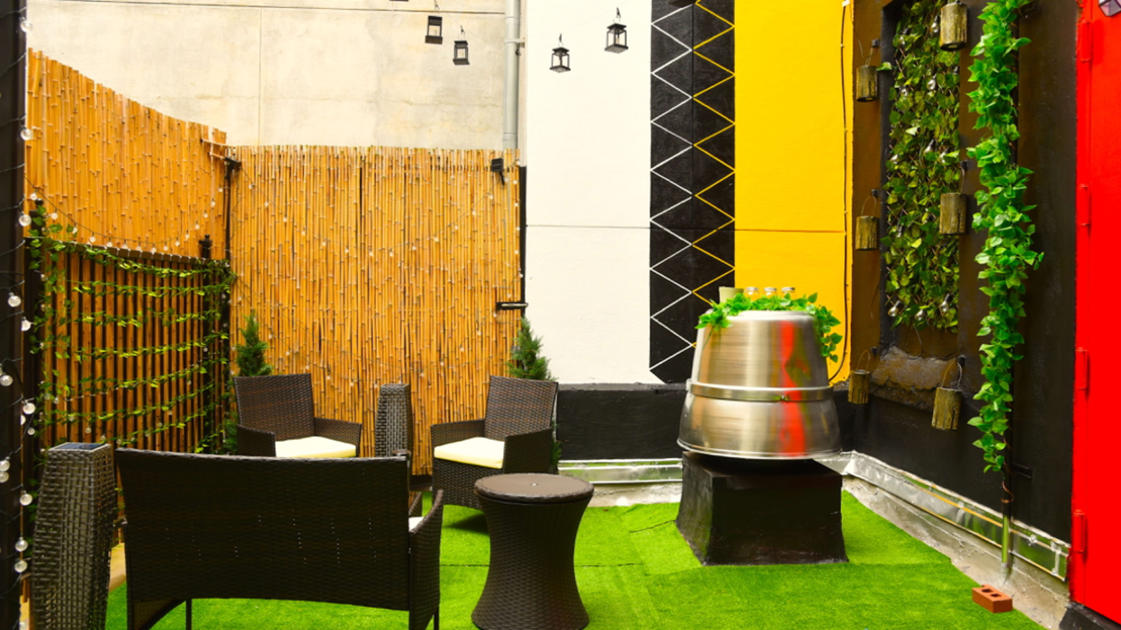 A small outdoor patio area with black wicker furniture, a metallic barrel, and green artificial grass surrounded by bamboo fencing and decorative wall elements.